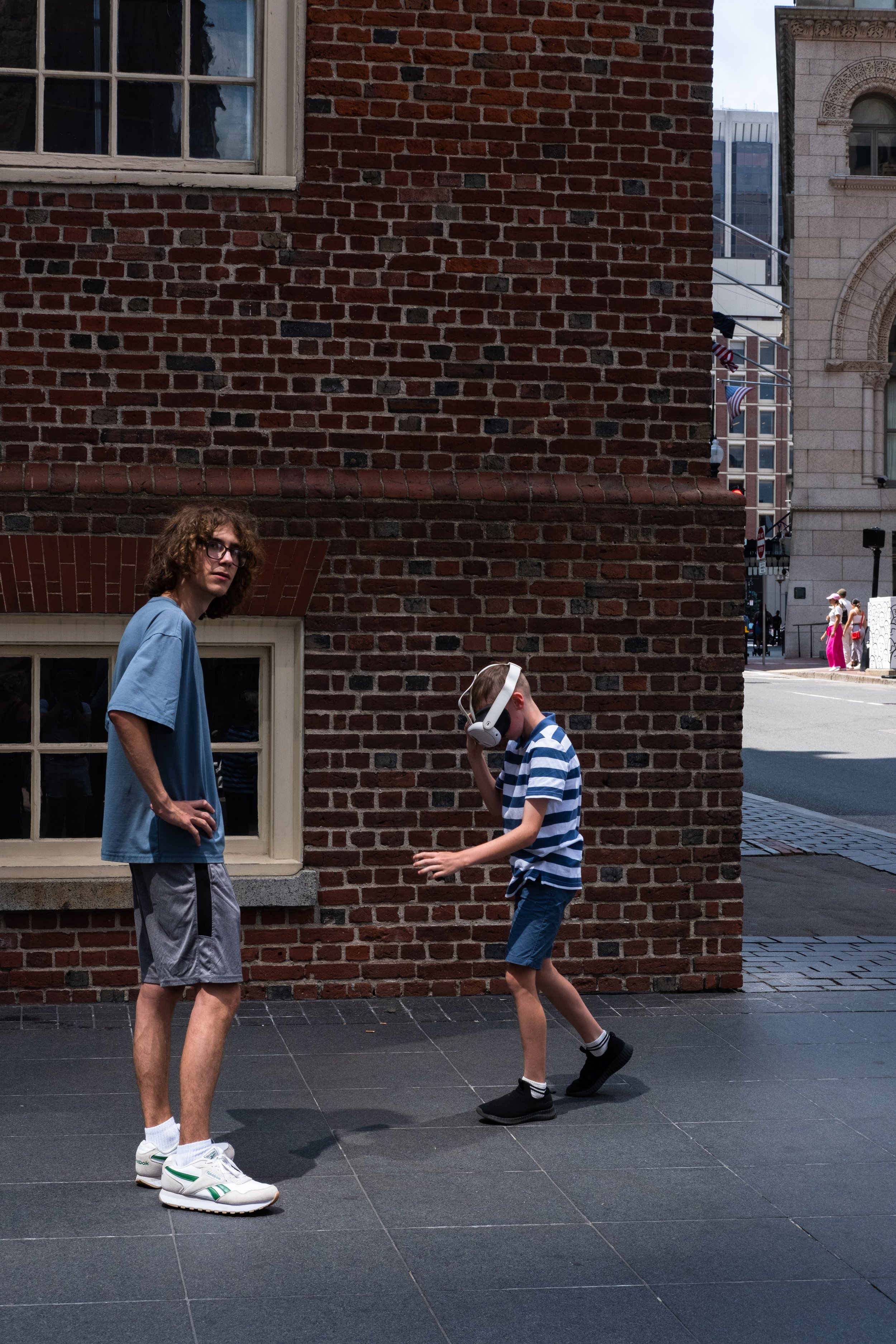 A young boy wearing a striped shirt, shorts, and black sneakers wearing a virtual reality headset, standing on a city sidewalk with a man in a blue shirt and shorts watching him. The background features a brick building with windows and part of the c