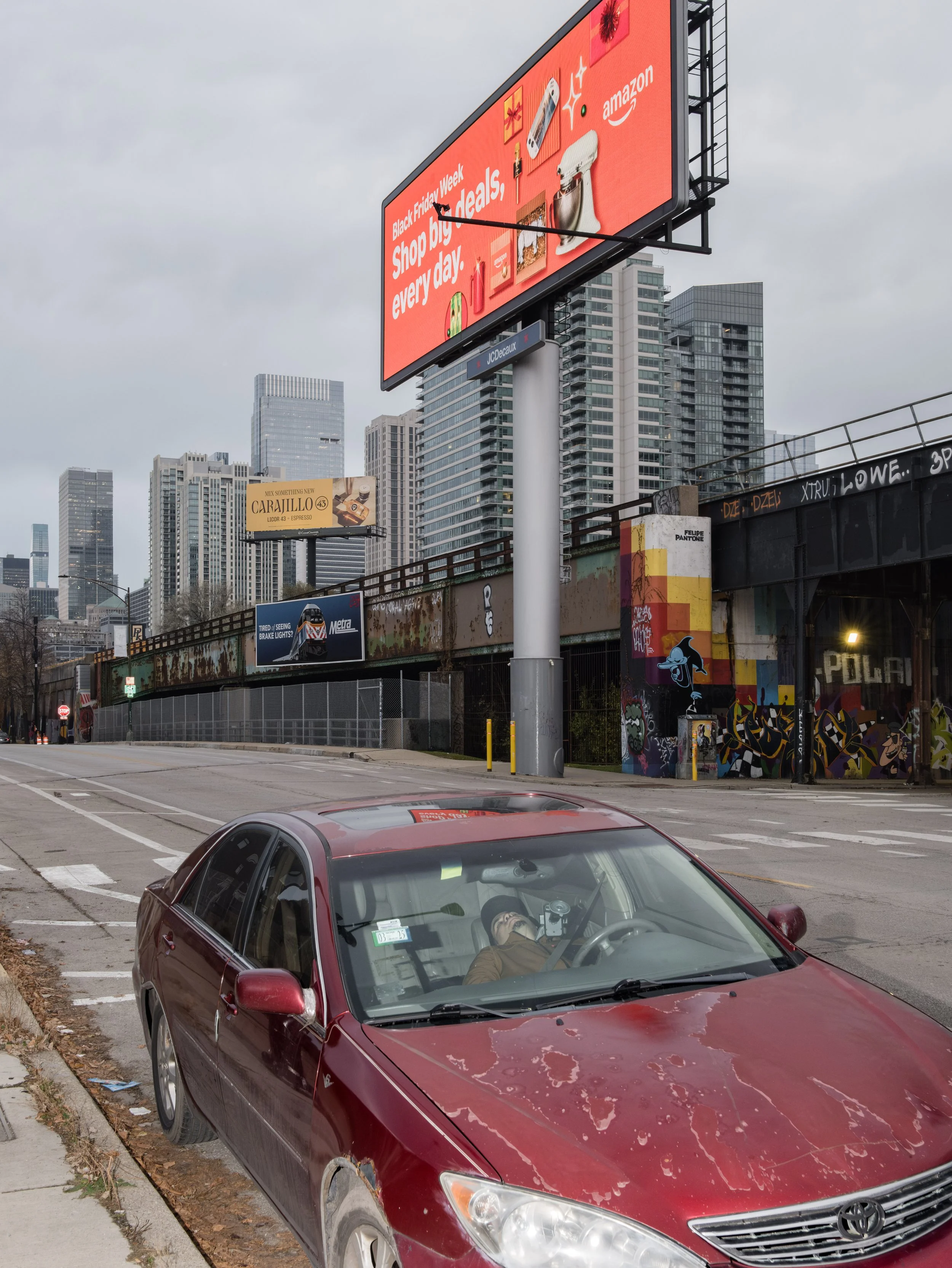A red sedan parked on the side of a city street with graffiti on the bridge and tall skyscrapers in the background. A person is sitting in the driver's seat wearing a mask and taking a photo with a camera through the windshield. Large digital billboa