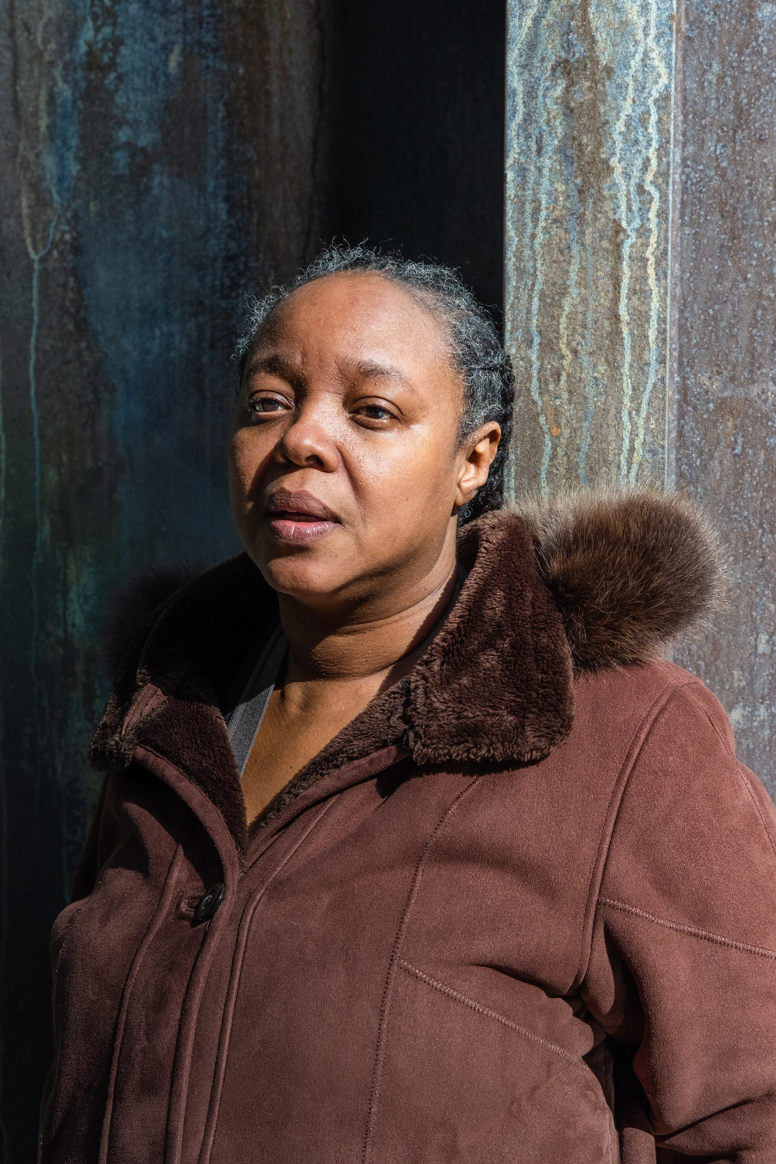 A woman with dark skin, wearing a brown coat with a fur-lined hood, standing in front of a textured, rust-colored wall.