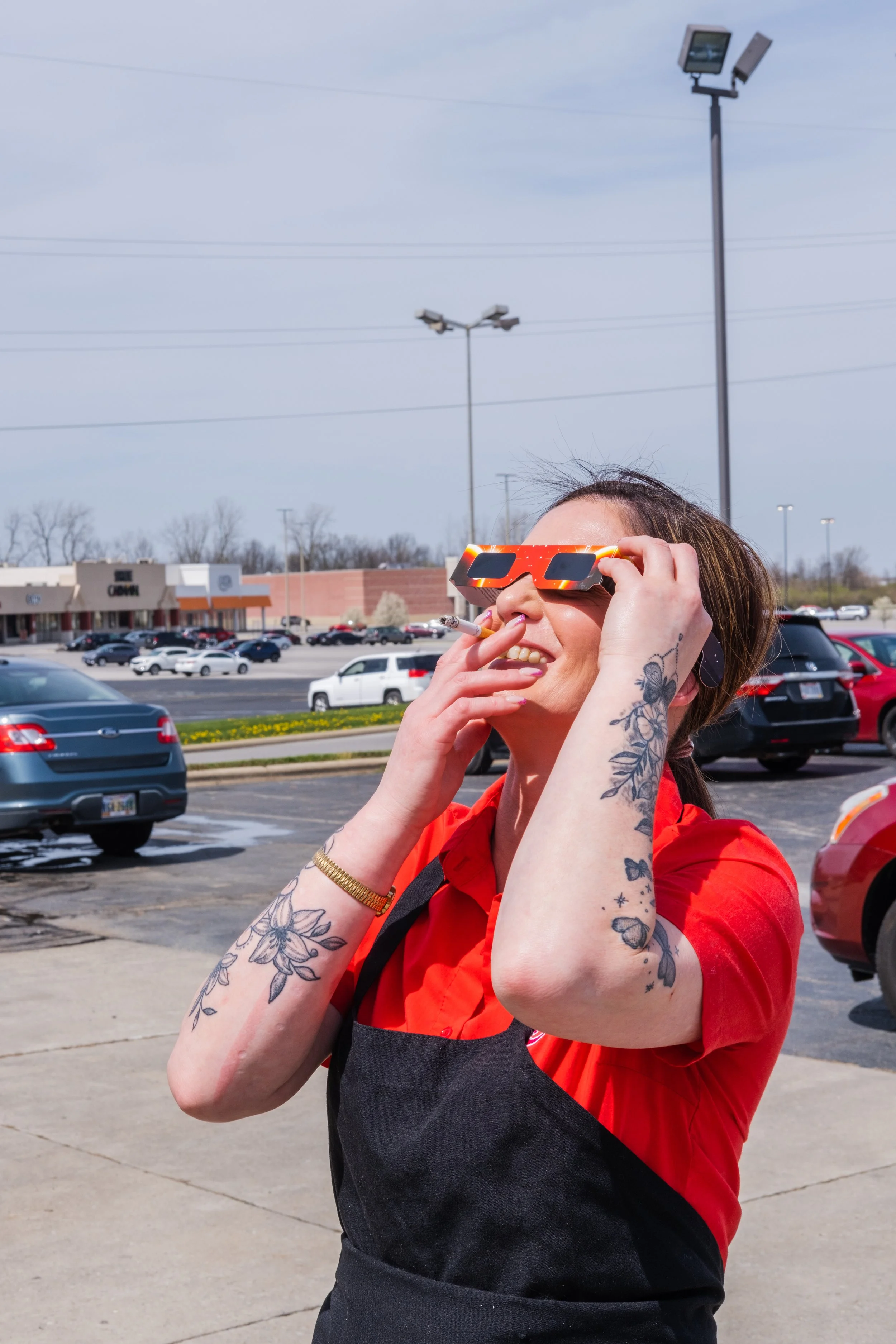 Smiling woman wearing orange sunglasses, a red shirt, and a black apron, smoking a cigarette in a parking lot on a sunny day.