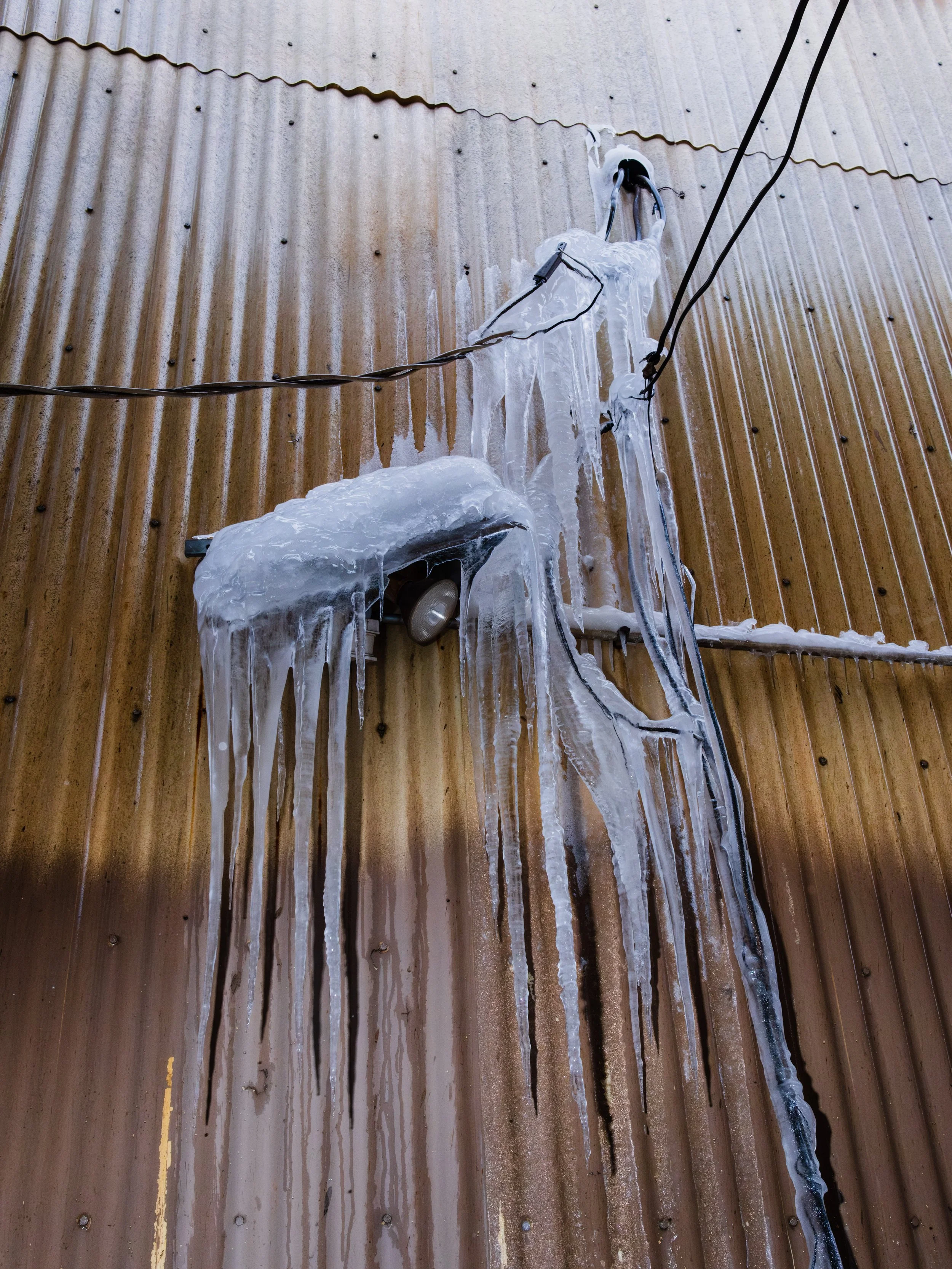 A brown wooden wall covered with icicles and ice formations around an electric meter and electrical wires.