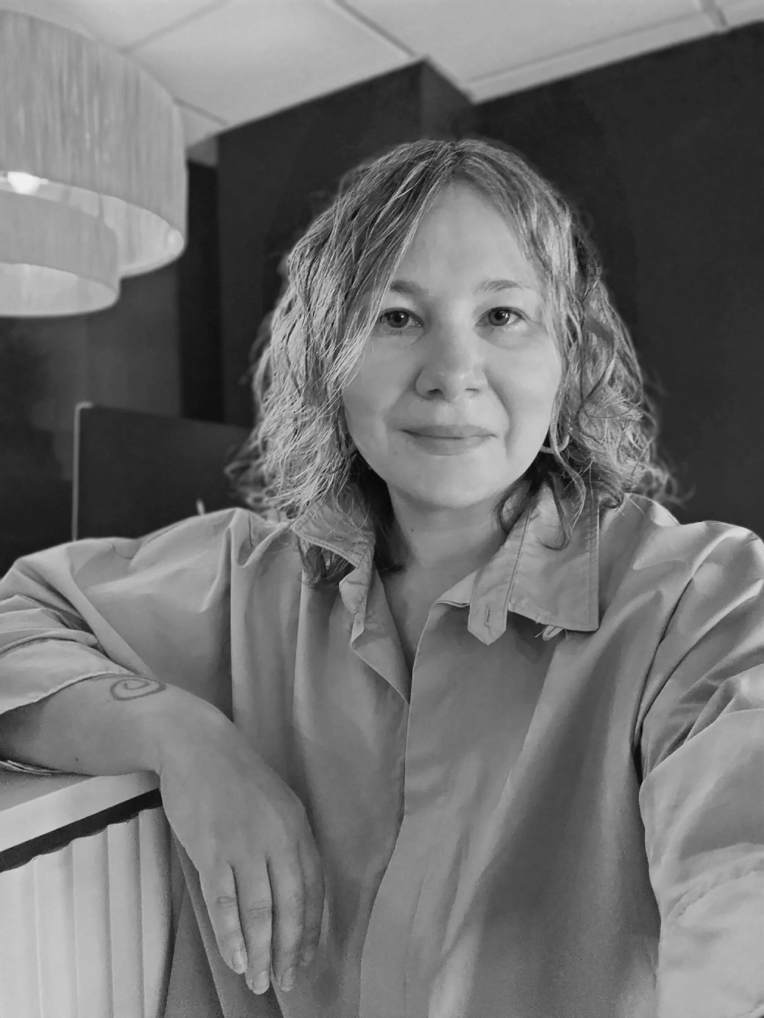 Black and white photo of a woman with wavy hair, wearing a button-up shirt, sitting with her arms crossed on a table, in an indoor setting with a lamp and dark wall in the background.