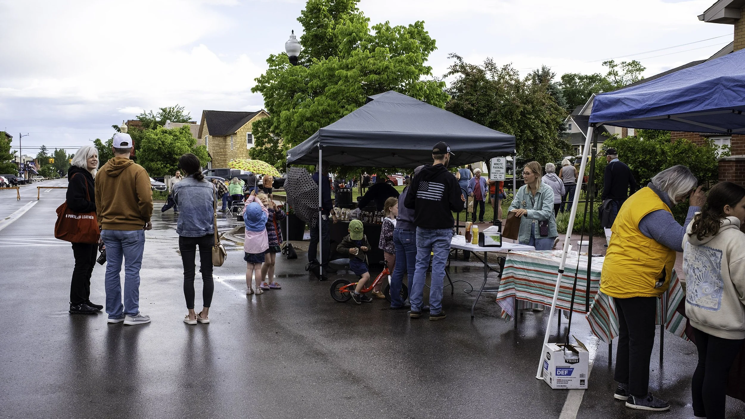 People at the Norway Michigan Open Air Market on a rainy day, with tents, tables, and shoppers browsing items. Some people are talking or looking at the vendor tables, and children are present.