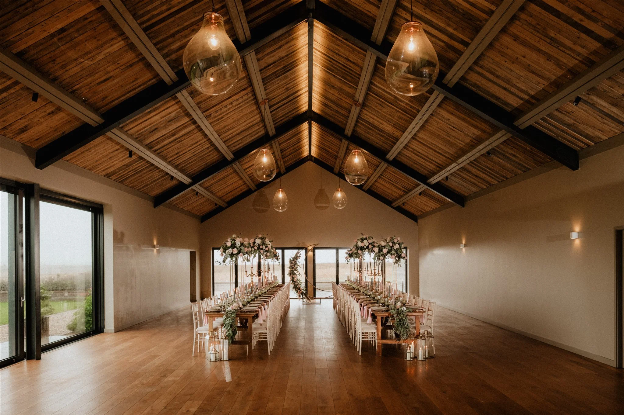 Wedding ceremony venue with two long tables, flowers, candles, and a decorated arch at the front, inside a spacious room with wooden ceiling and floor.