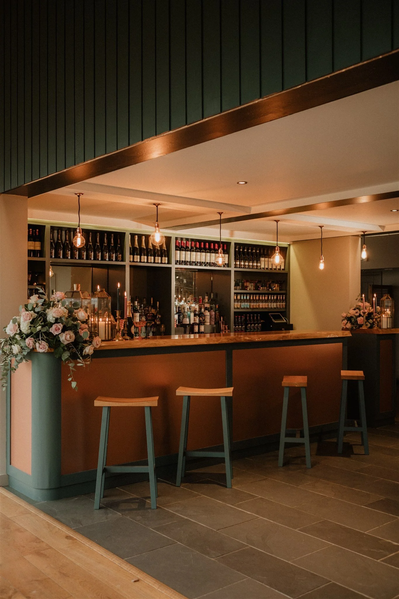 A cozy bar with a wooden counter, floral arrangements, and warm lighting with exposed bulbs.