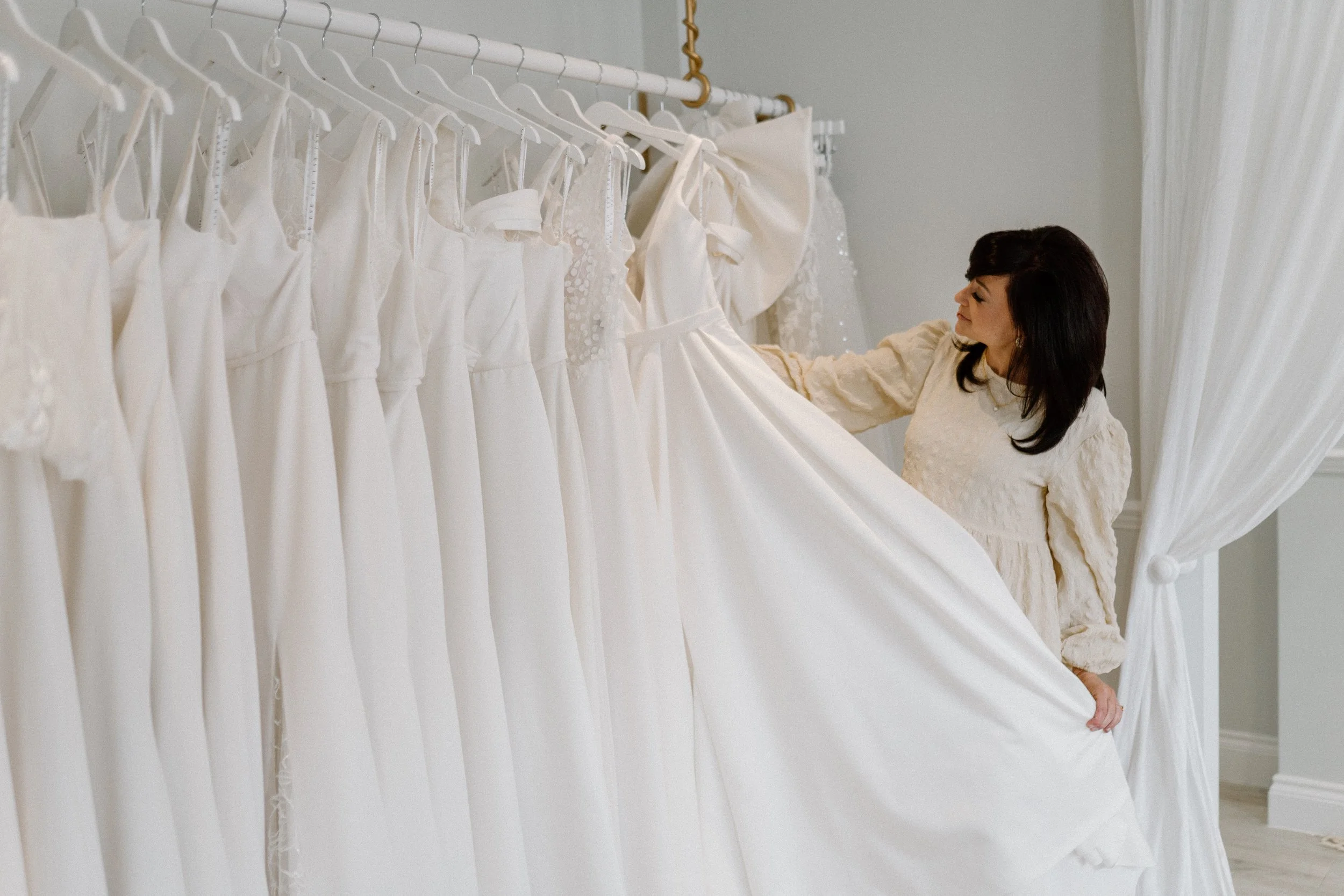 A woman in a beige dress shopping for wedding dresses, looking at a row of white wedding gowns hanging on a rack.