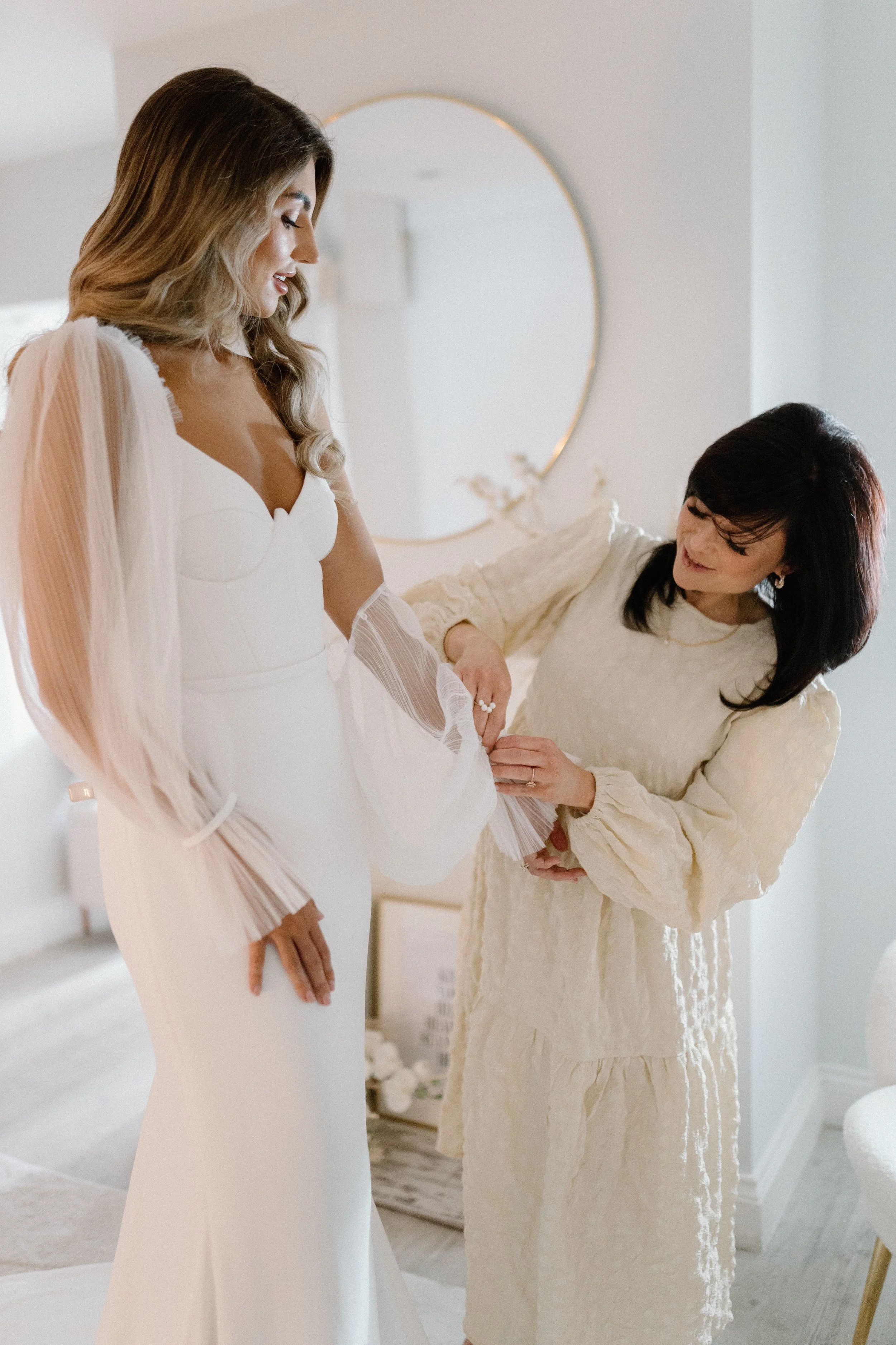 A bride in a white wedding dress with sheer long sleeves is assisted by a woman in a cream-colored dress, likely helping her with her dress or accessories in a softly lit room with a circular mirror on the wall.