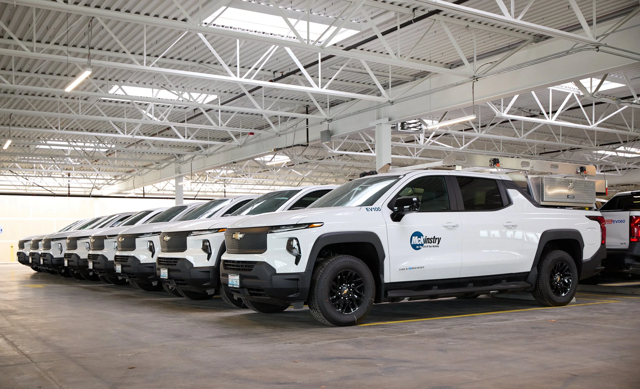 8 white pick up trucks lined up side by side inside a warehouse. The trucks have the McKinstry logo printed on the front driver-side door.