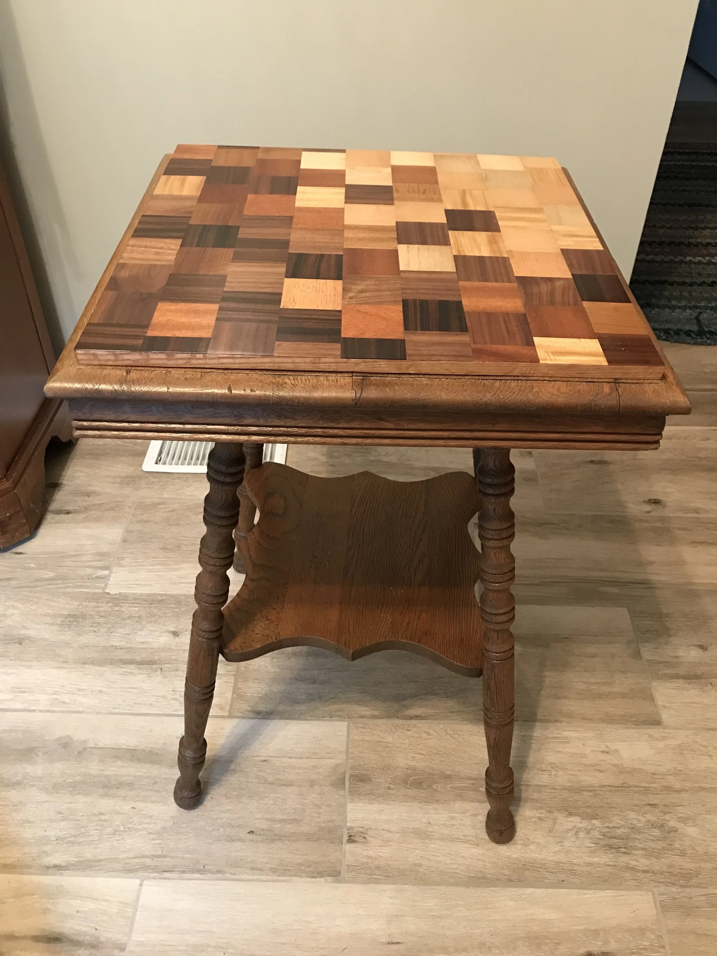 Wooden side table with a checkered mosaic top, featuring various shades of brown and beige wood, and ornate turned legs on a wooden floor.