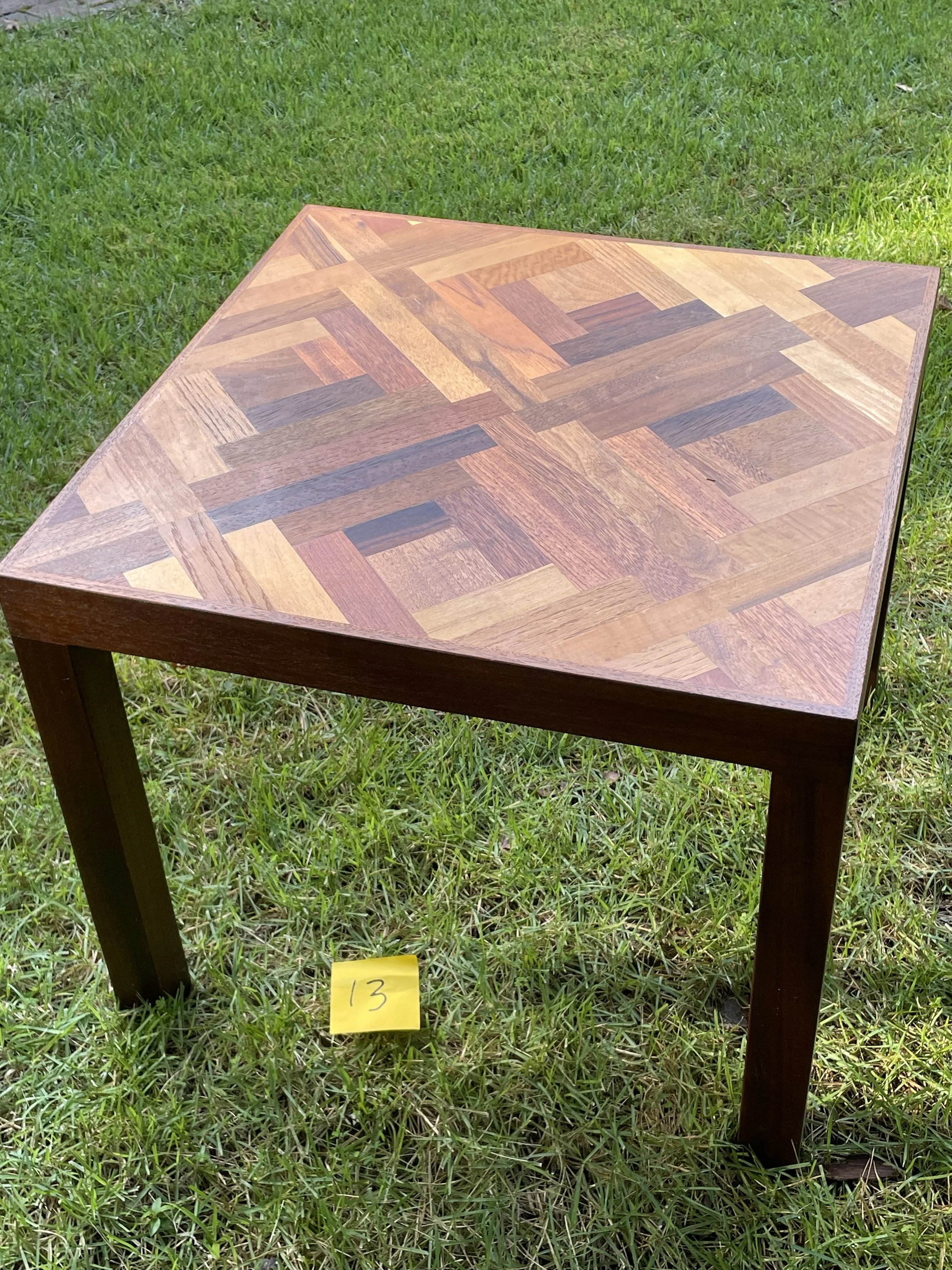 A small square wooden table with a geometric patterned top, placed on grass with a yellow sticky note marked '13' nearby.