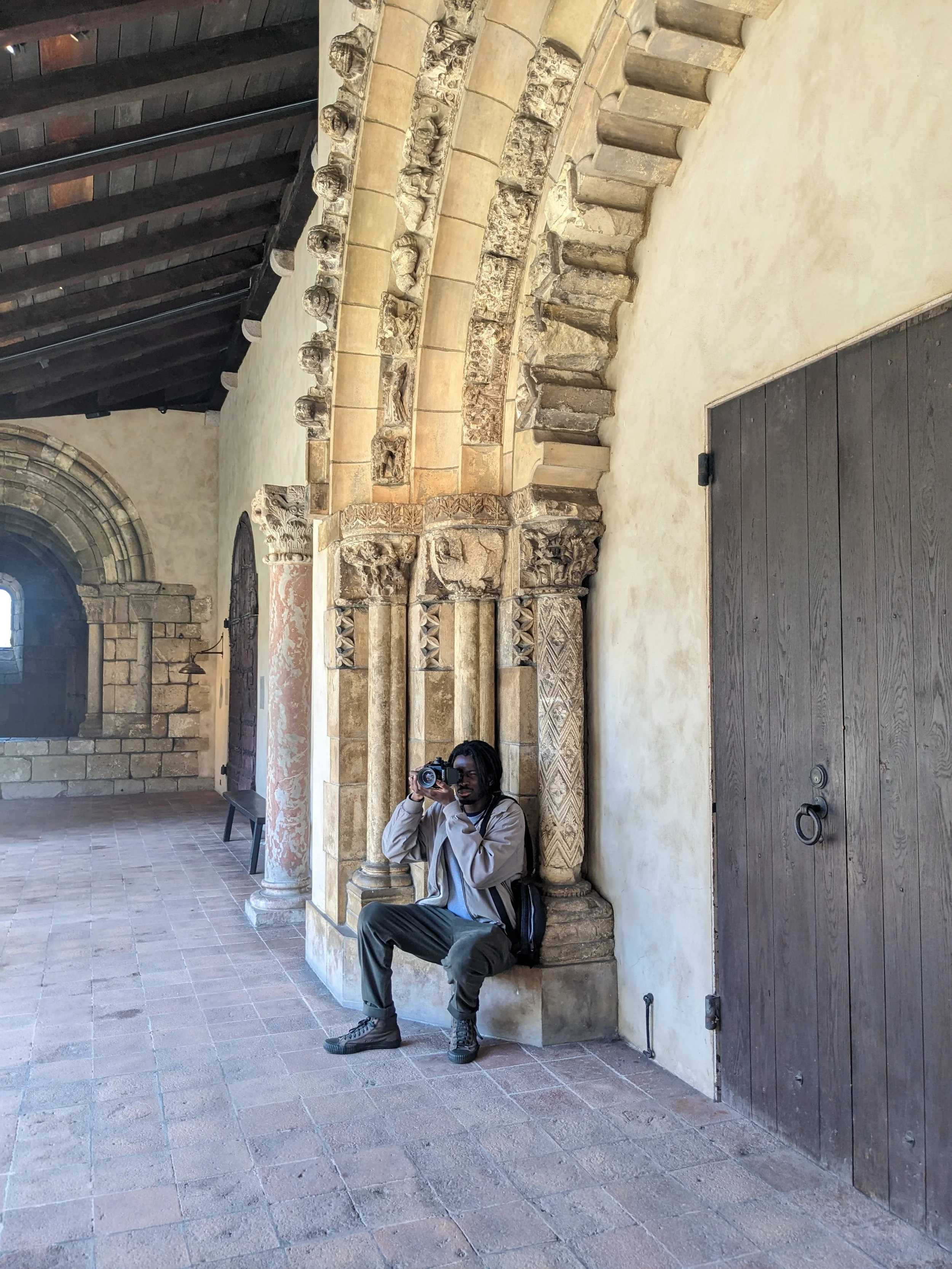 Person sitting on a stone base of ornate architectural column, taking a photo with a camera in a historic building with stone and wood features.