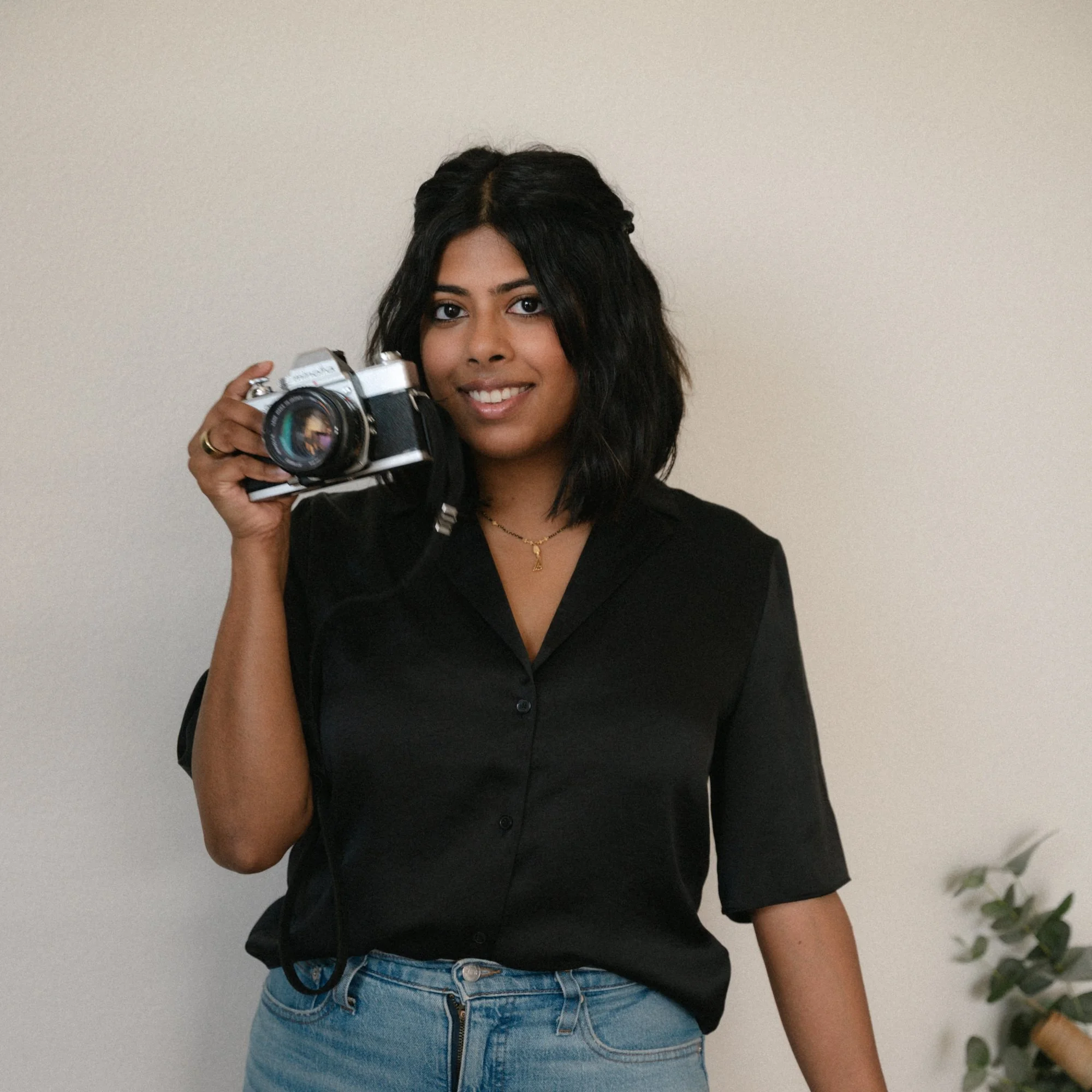 A woman with black wavy hair, wearing a black button-up shirt and blue jeans, holding a vintage film camera, smiling at the camera, standing against a plain off-white wall with a small plant visible on the right side.