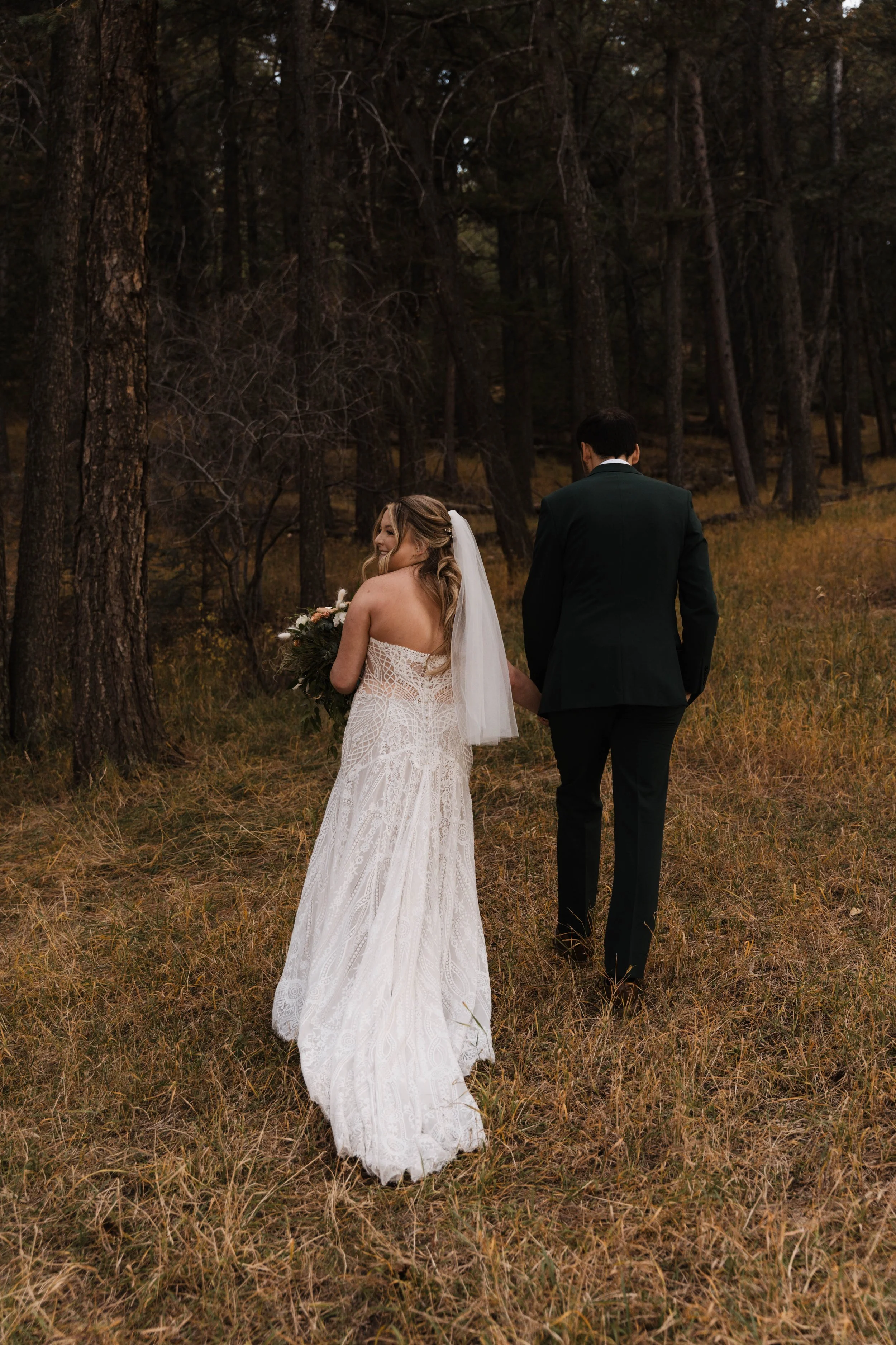 Bride and groom holding hands walking through a forest, bride wearing a white lace wedding dress and veil, groom in a dark suit, tall trees in background.