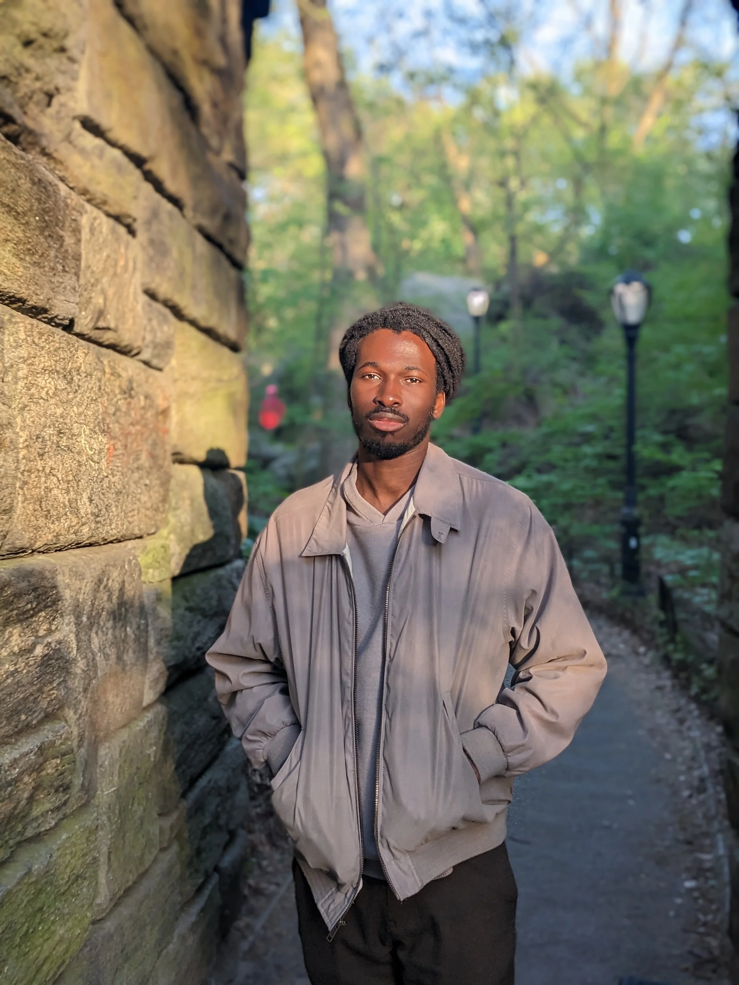 A man stands with hands in pockets outdoors next to a stone wall, with trees and street lamps in the background during daylight.