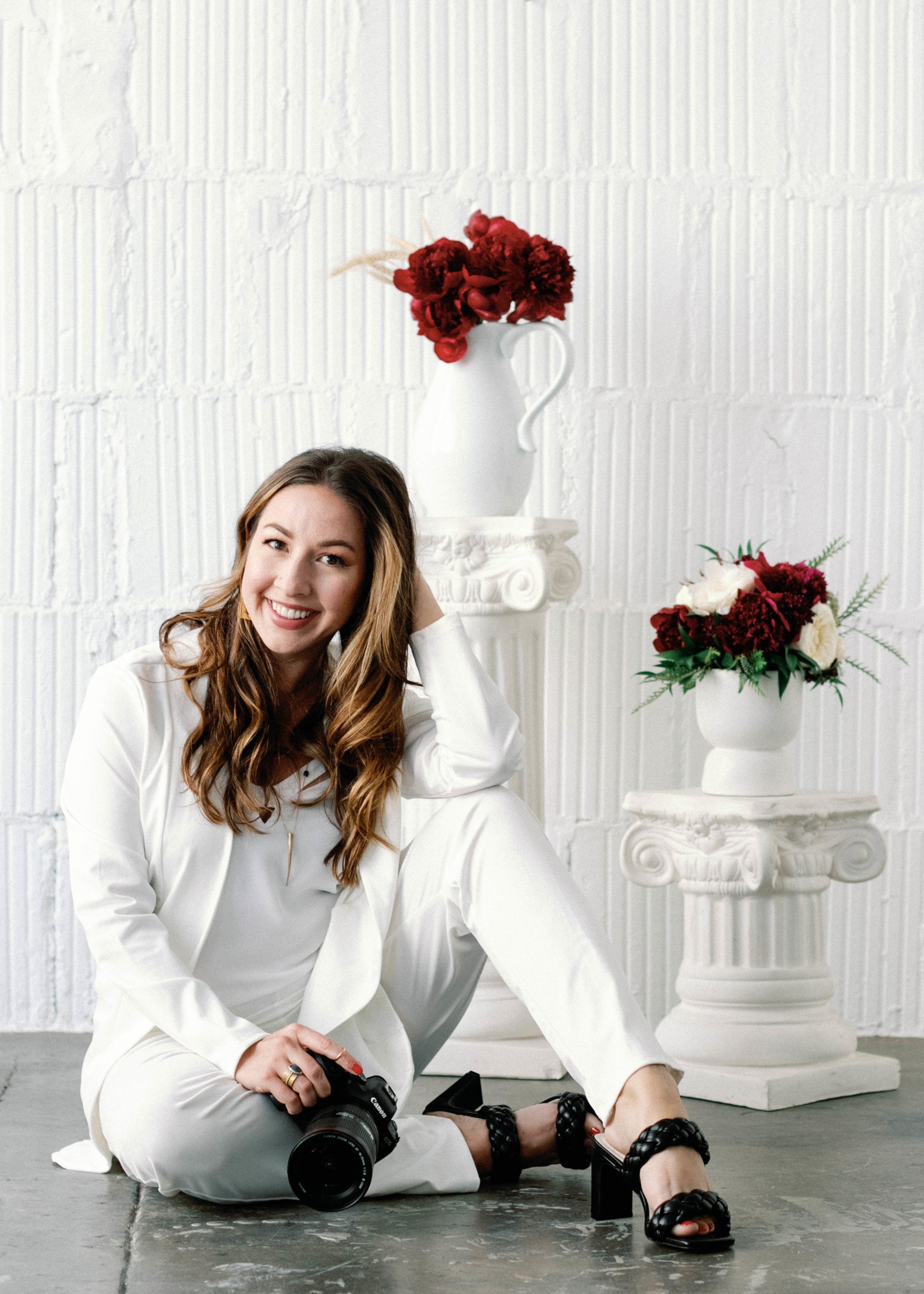 A woman with long wavy brown hair sitting on the floor in a white suit, holding a camera, and smiling at the camera. There are white vases with red and white flowers on white pedestal stands behind her, against a white brick wall.