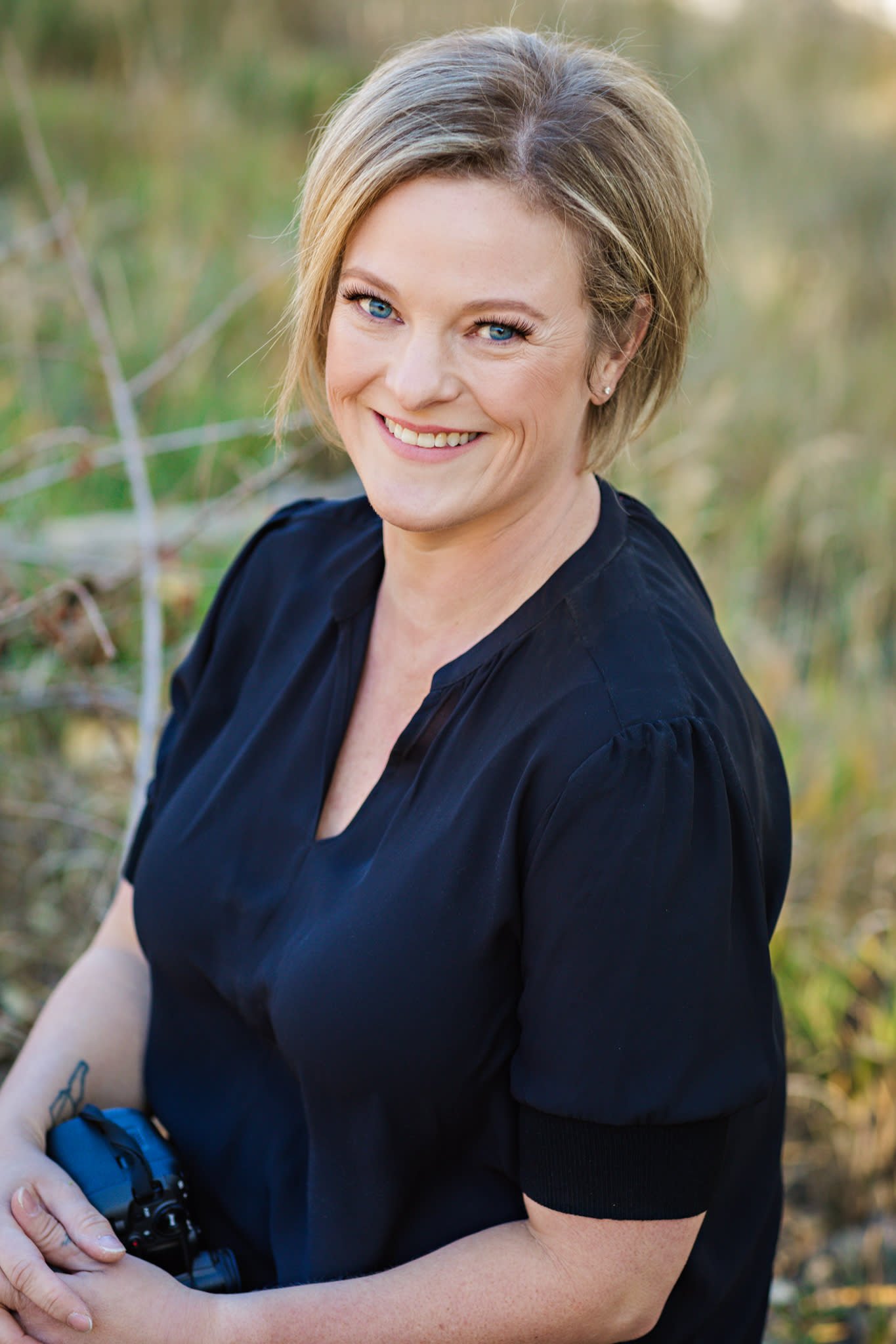 A woman with blonde hair, blue eyes, and a friendly smile sitting outdoors in a natural setting. She is wearing a black top and holding binoculars.