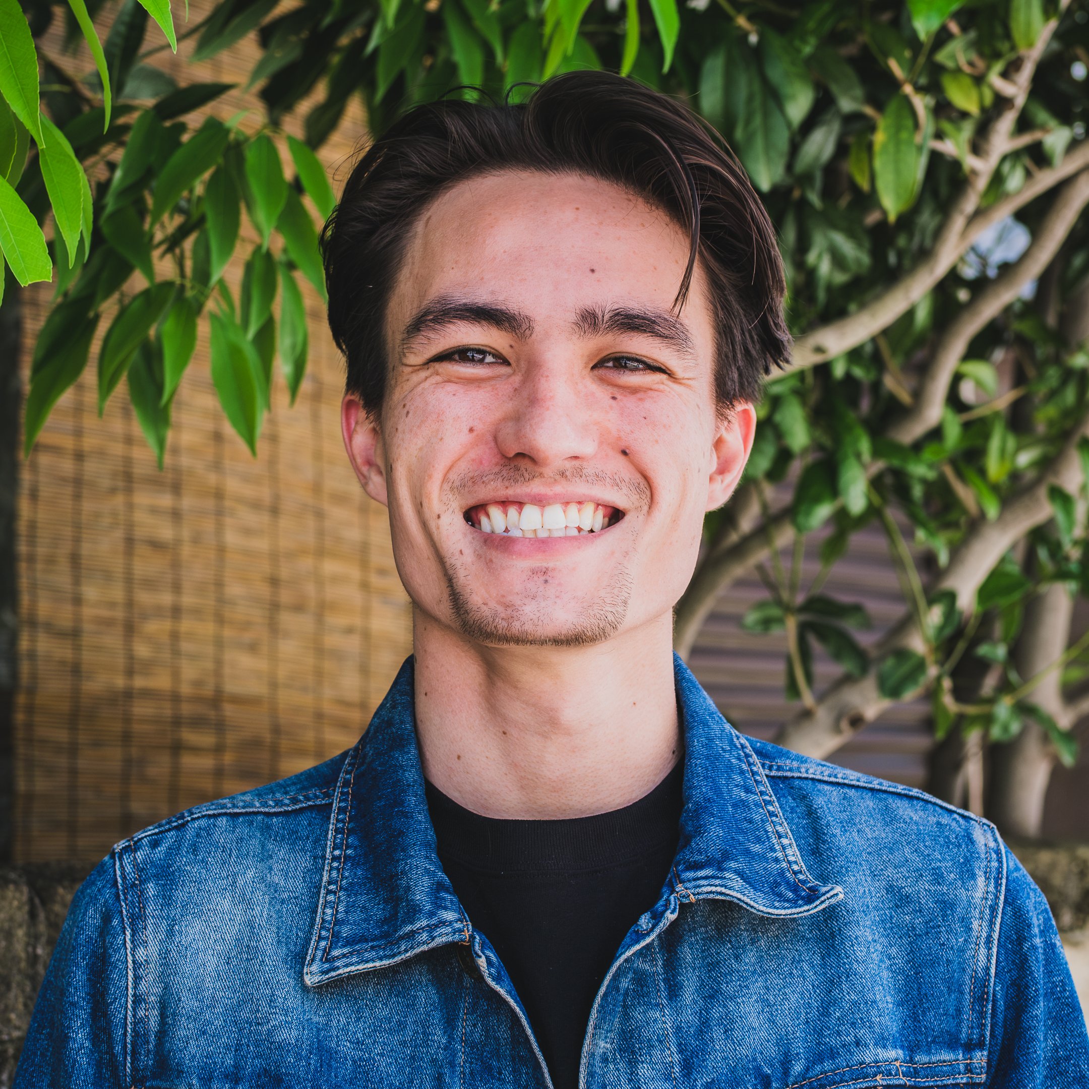 Smiling young man with dark hair, wearing a denim jacket and black shirt, standing outdoors in front of green leafy plants.