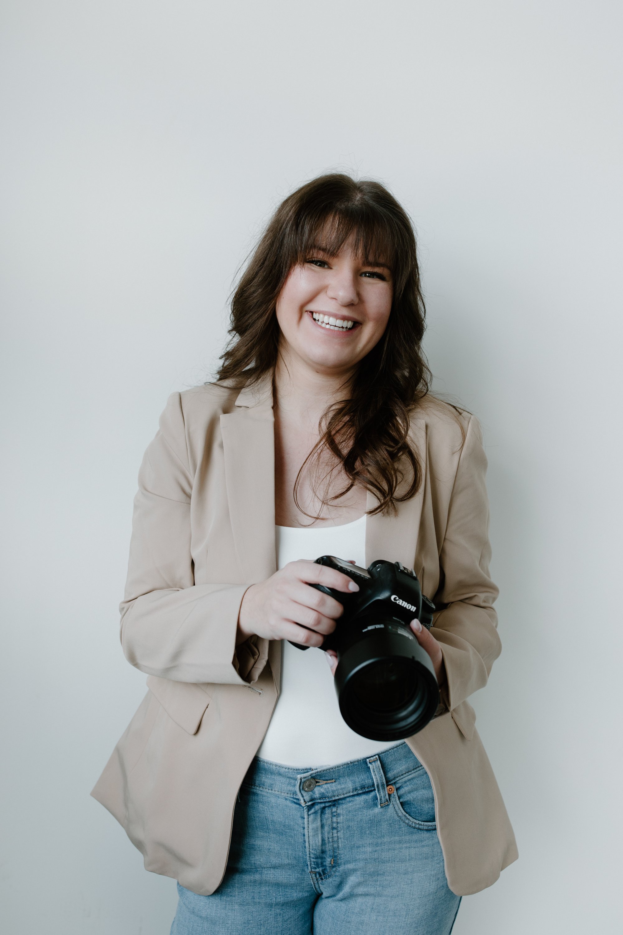A woman with brown hair smiling and holding a Canon DSLR camera, standing against a plain off-white background.