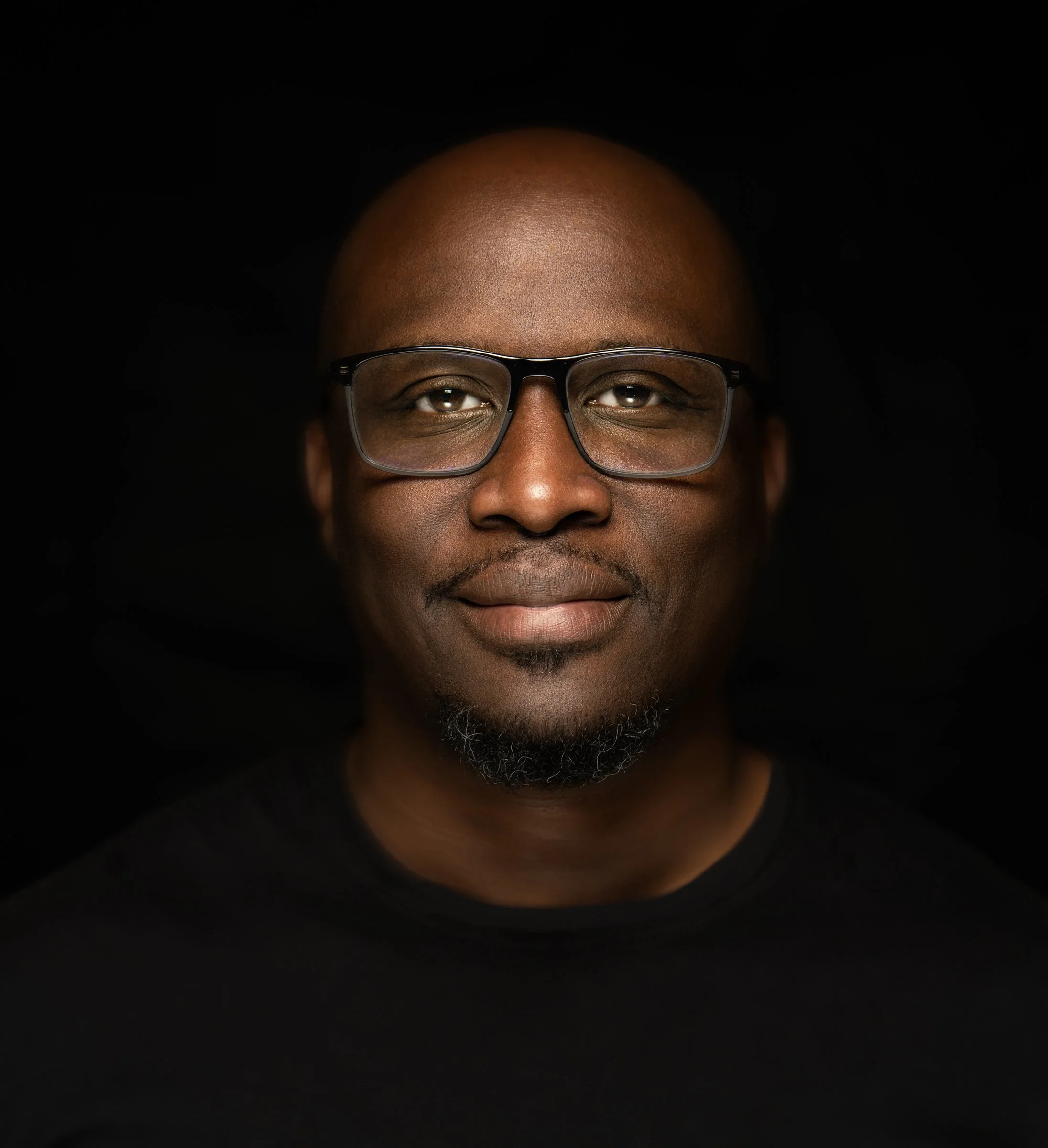 Portrait of a man with dark skin, wearing glasses, looking directly at the camera, against a black background.