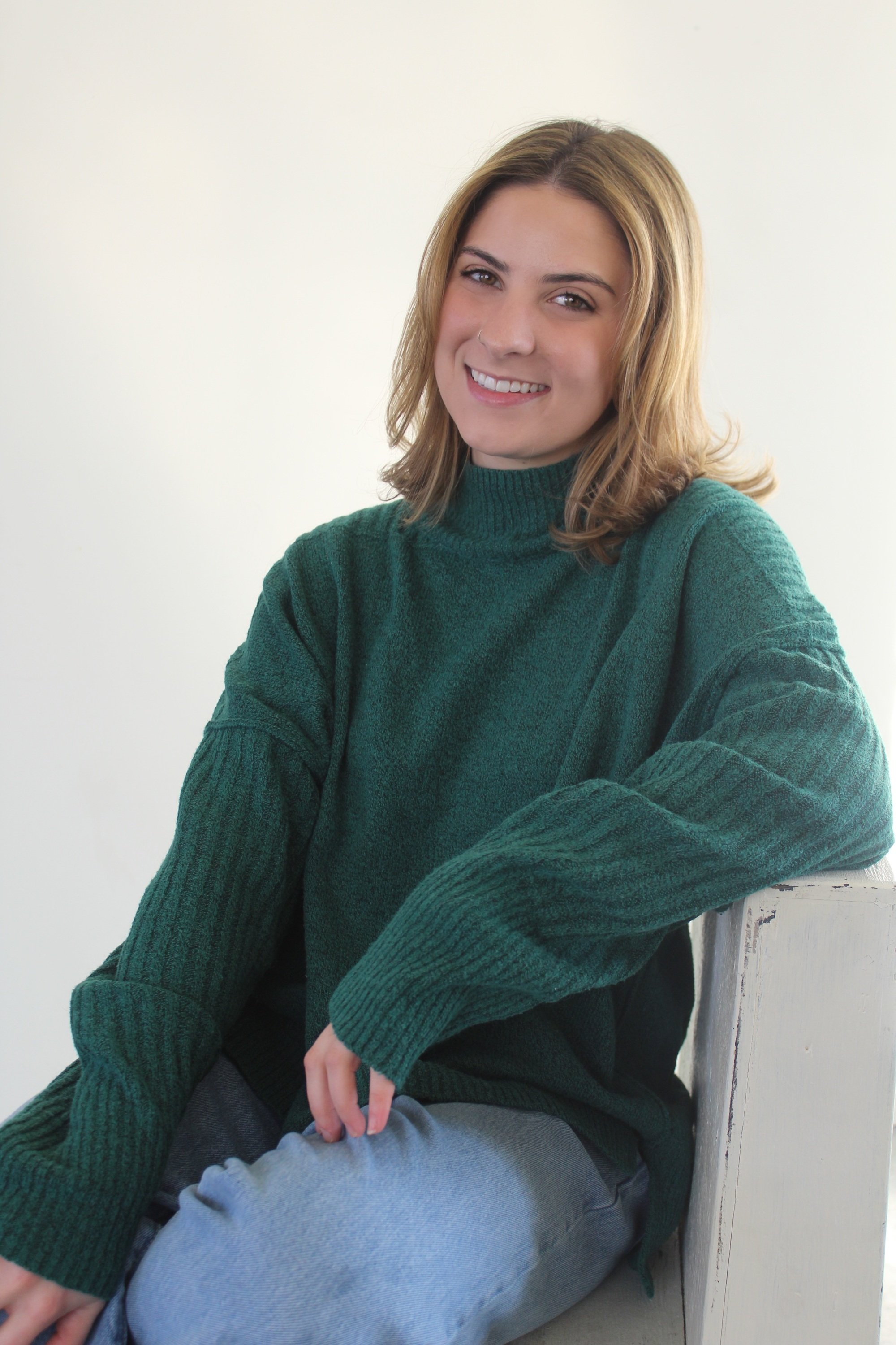 A young woman with shoulder-length blonde hair, smiling, wearing a green sweater and light gray pants, sitting on a white wooden surface against a plain white background.