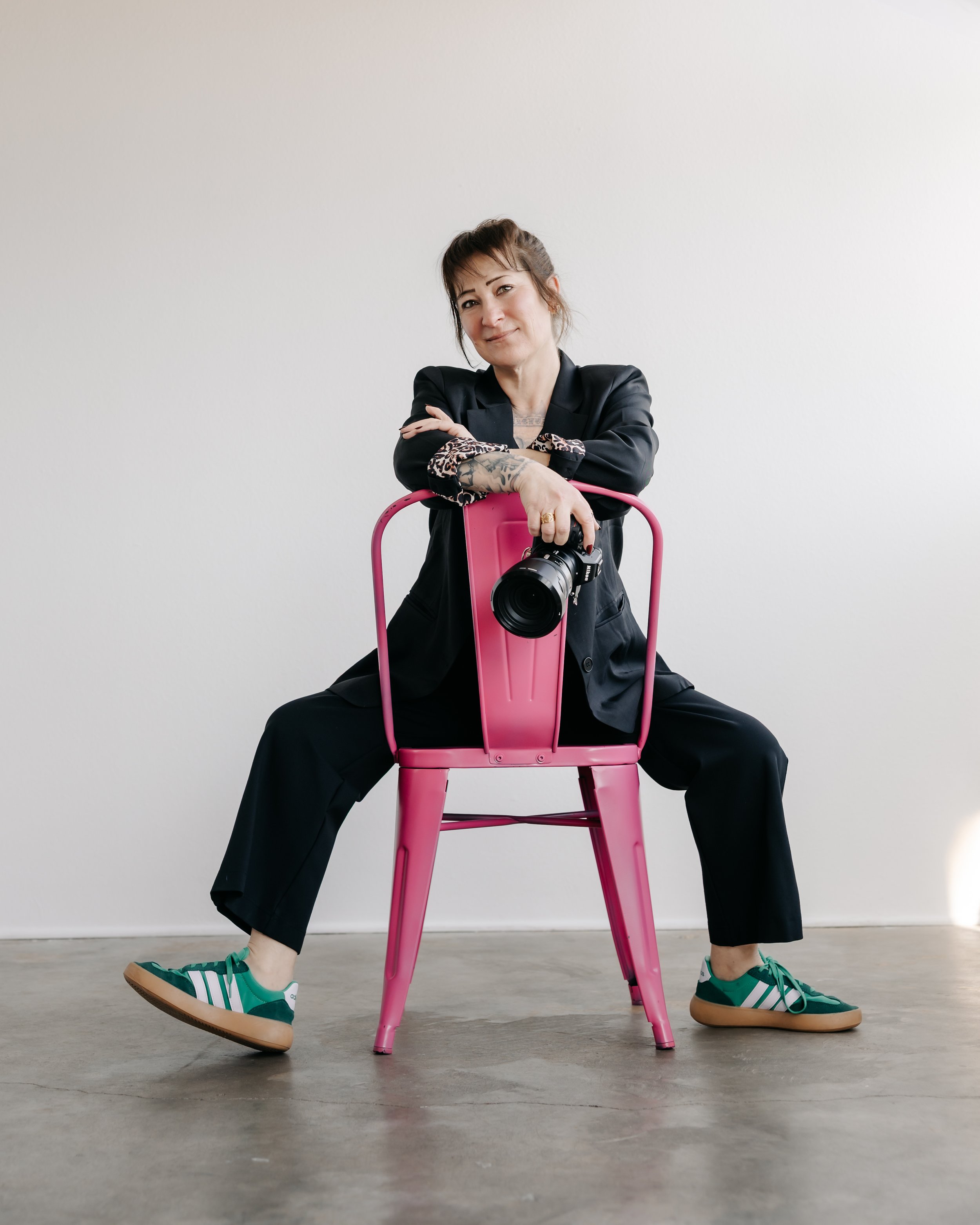 A woman in a black blazer and black pants is sitting on a pink chair, holding a camera, with one arm crossed over her chest. She is wearing green and white sneakers and is posed with her legs wide apart against a plain white wall.