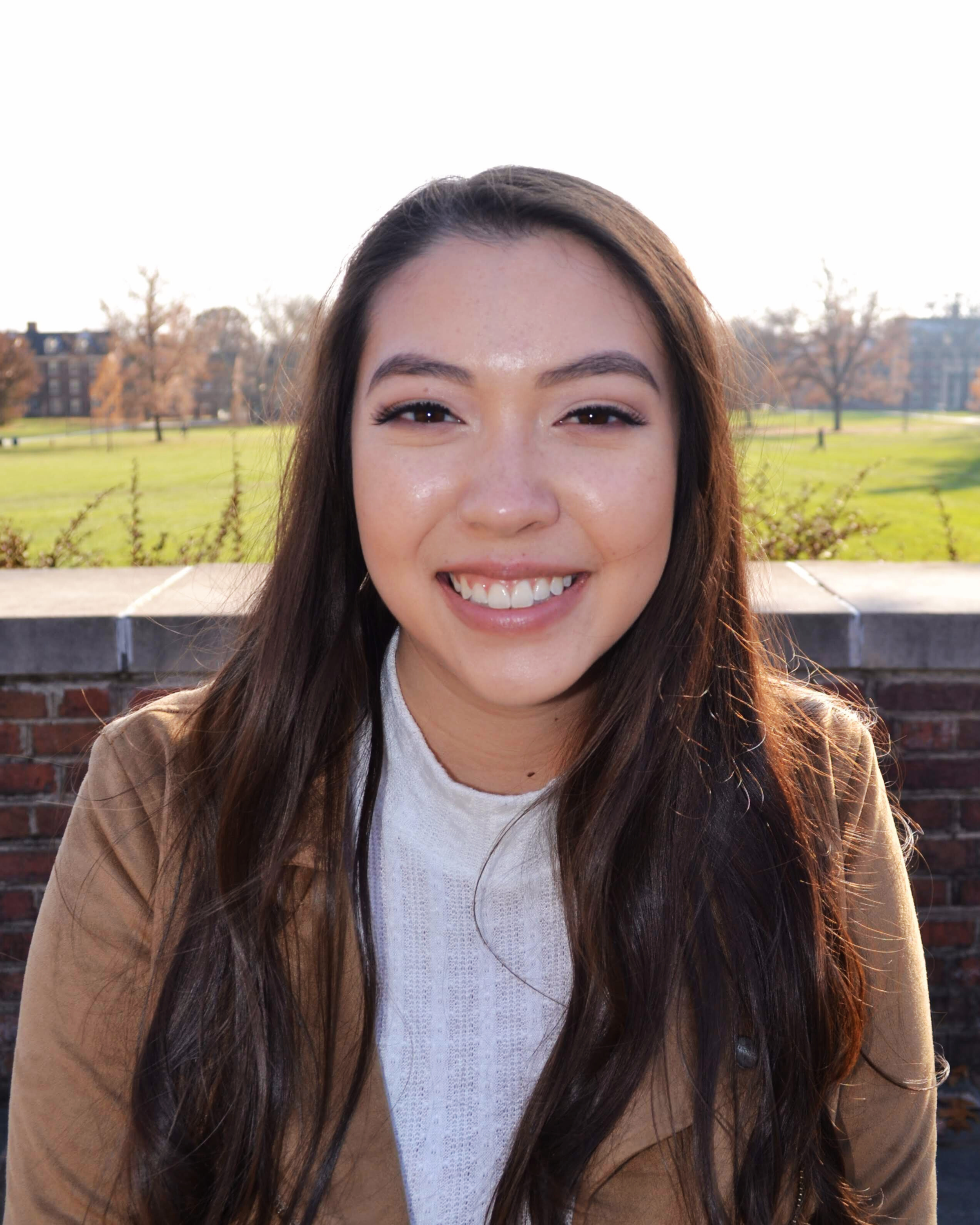 A young woman smiling outdoors with a park and trees in the background during daytime.