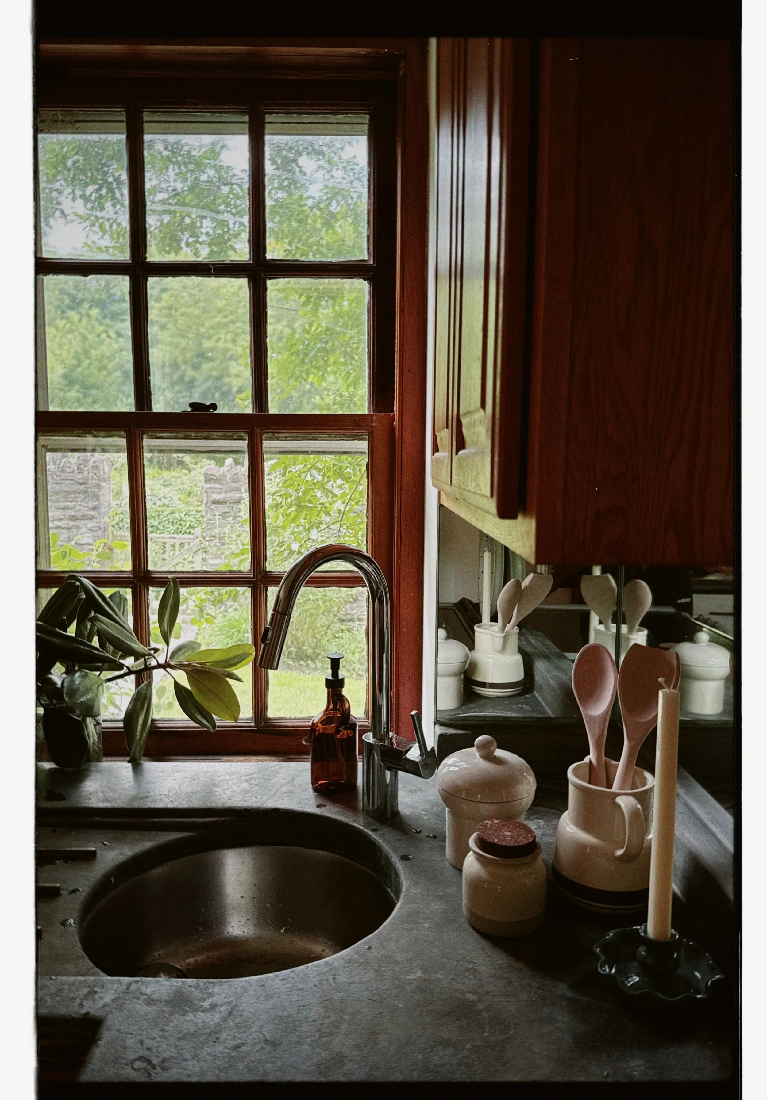 A kitchen sink area with a wooden window behind it, a plant to the left, and various cooking utensils and containers on the countertop.