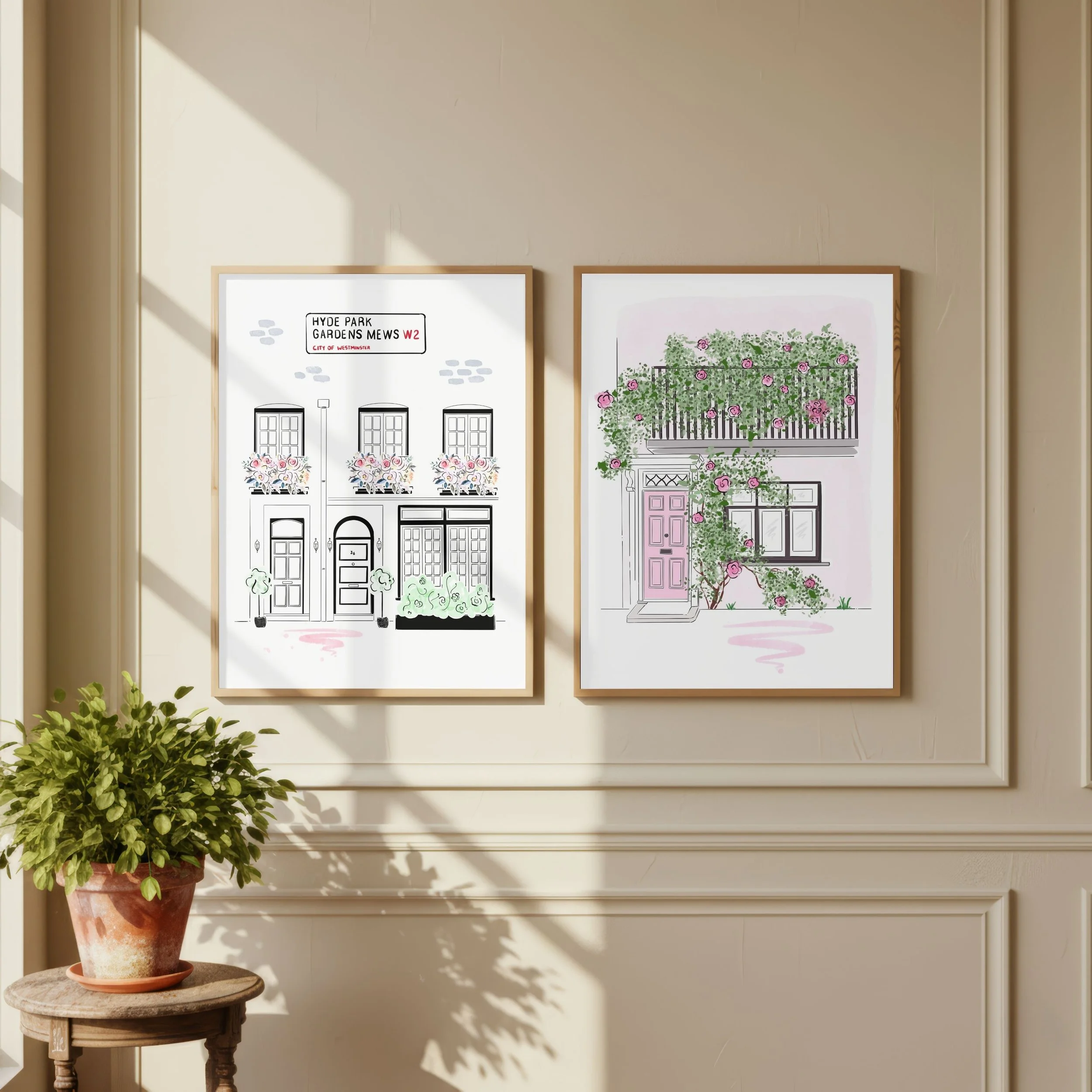 Two framed pictures of house facades with floral and greenery designs on a beige wall, with a potted plant on a small round wooden table in front, casting shadows on the wall.
