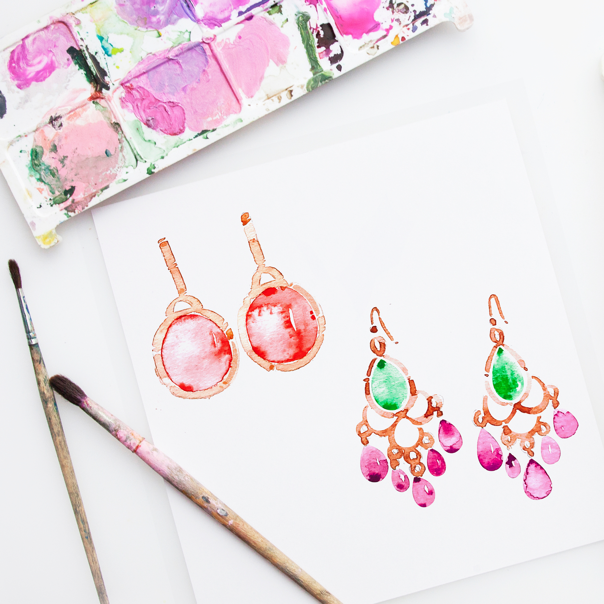 A watercolor painting of earrings,, painted with shades of orange, pink, and red, with green ears, on a white sheet of paper, with a white background. A paintbrush with pink and purple paint is placed on the left side of the paper.