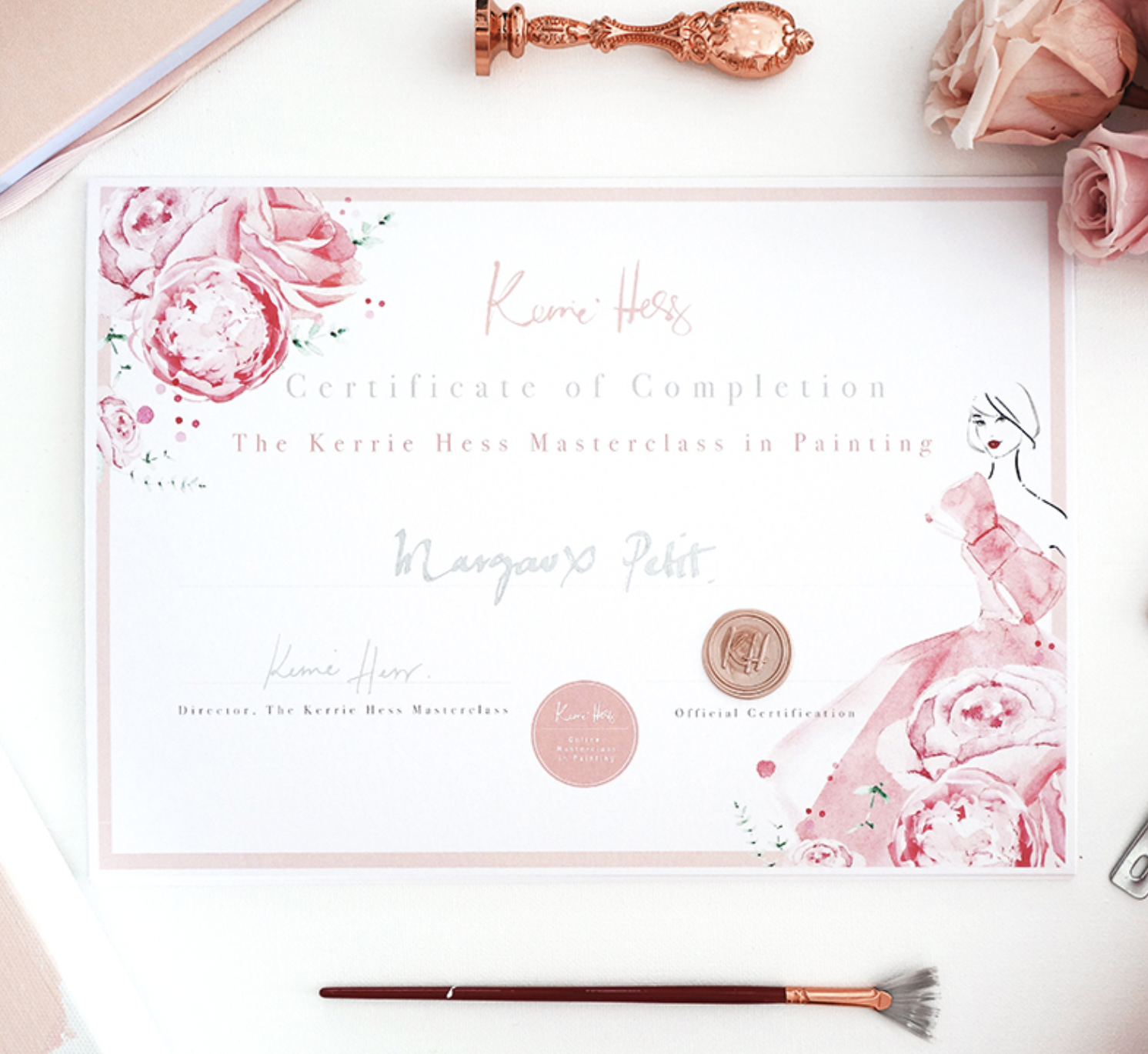 A certificate of completion for a painting masterclass by Kerrie Hess, decorated with pink watercolor roses and a drawing of a woman in a pink dress, placed on a desk with a rose gold wax seal, a pink decorative wax seal stamp, pink roses, and a pink and rose gold paintbrush.
