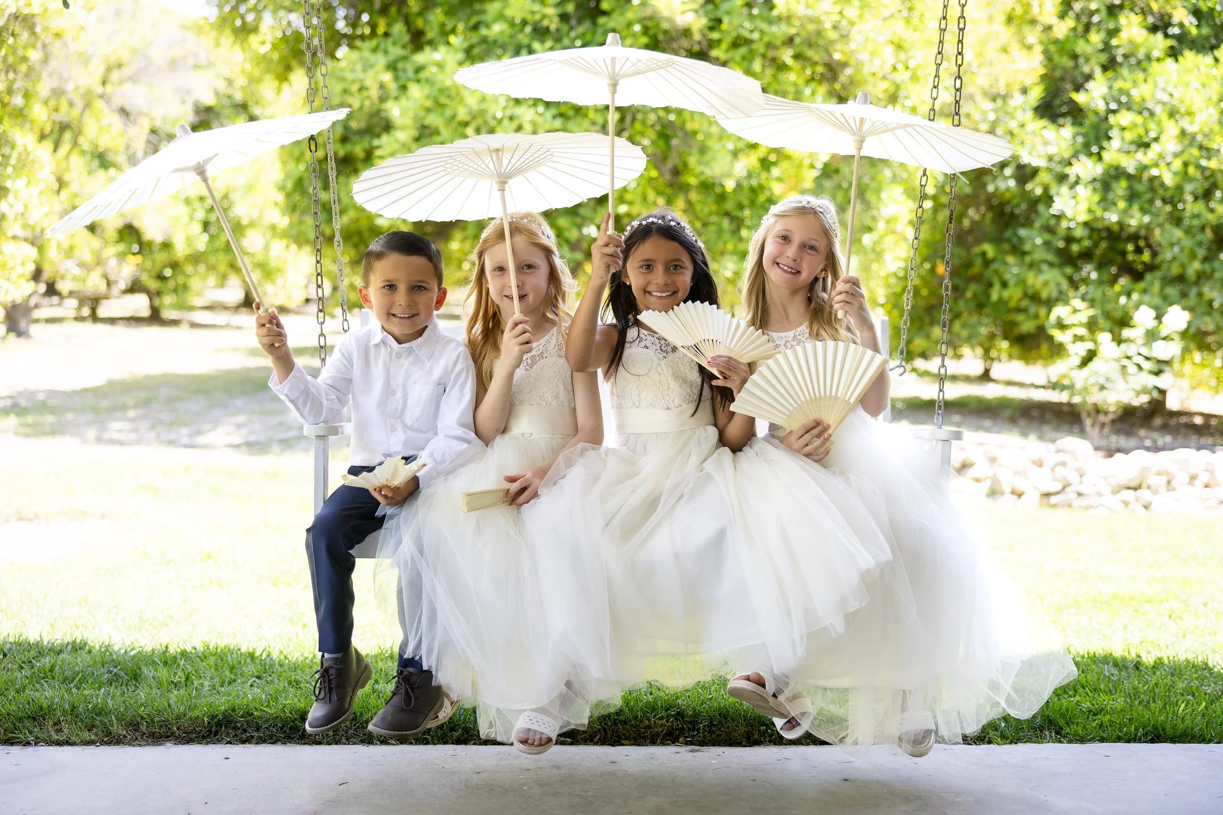 Four children, dressed in light-colored dresses and dark pants, sitting on a swing outdoors with green trees in the background. They are holding white umbrellas and paper fans, smiling at the camera.