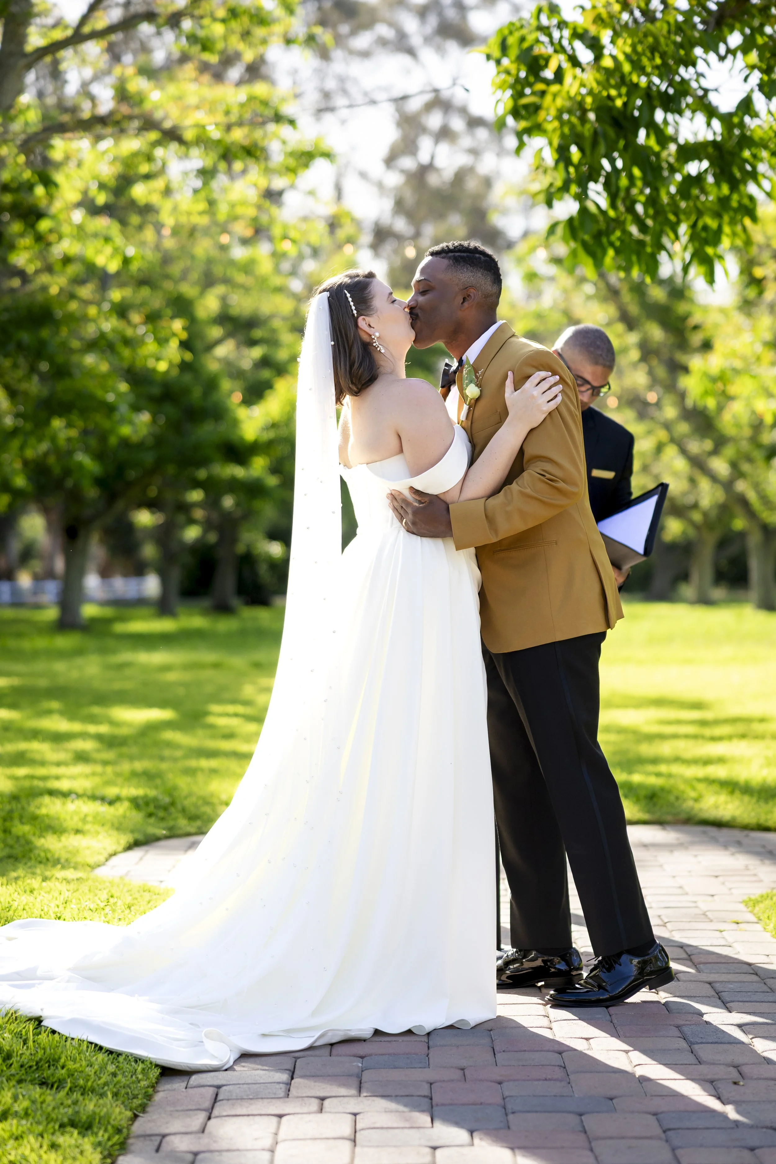 A bride and groom share a kiss during their outdoor wedding ceremony on a sunny day, with a officiant in the background holding a book and a folder, surrounded by green trees and grass.