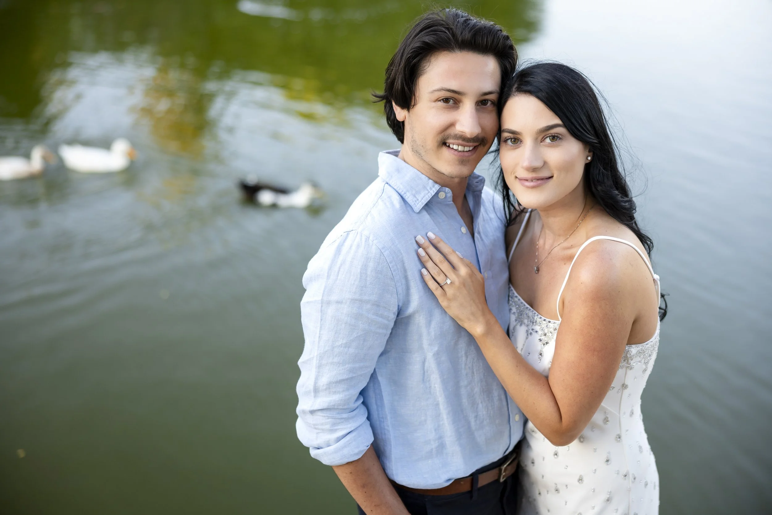 A couple standing close together outdoors by a body of water, smiling at the camera, with ducks swimming in the background.