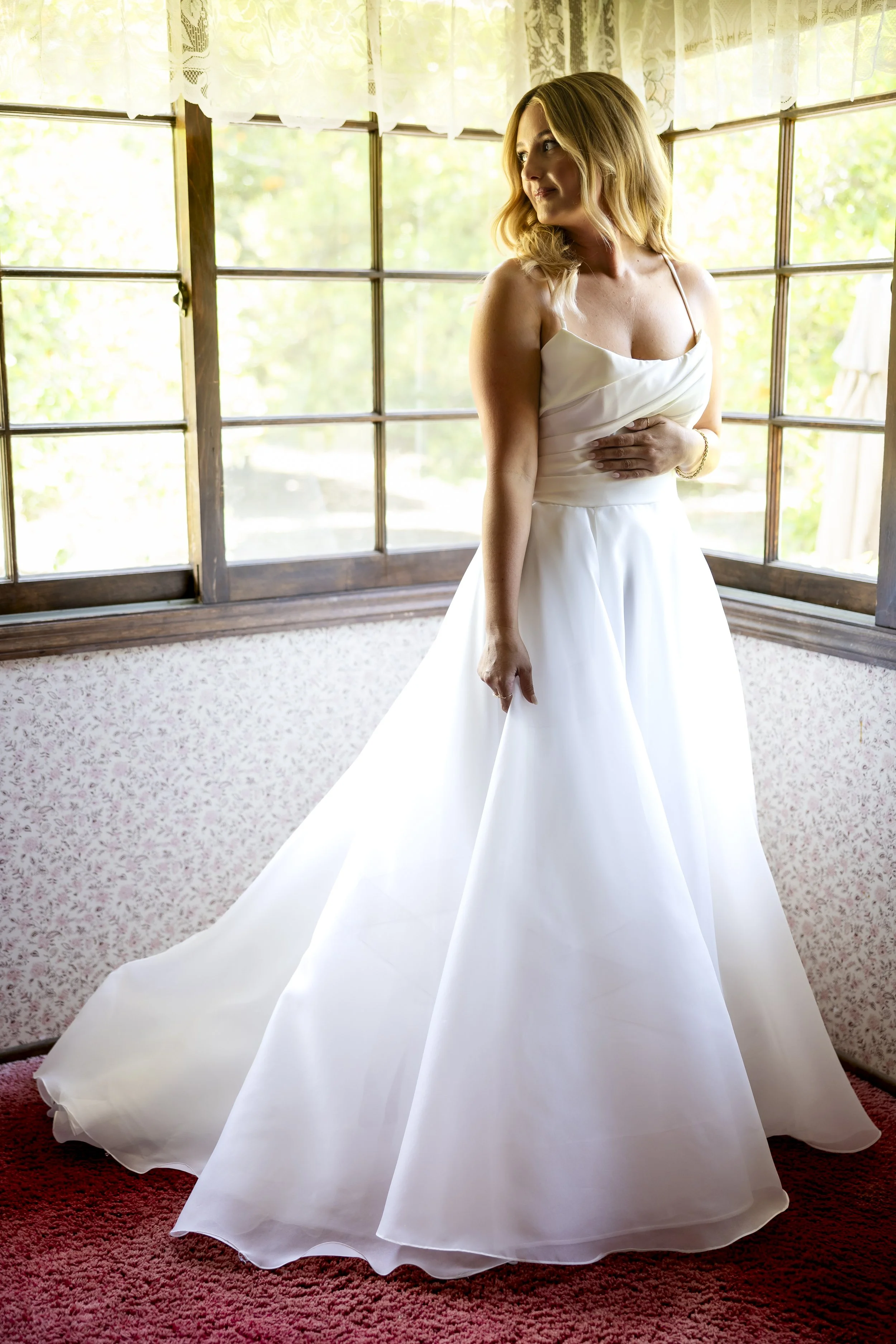 A woman in a white wedding dress standing indoors near large windows with lace curtains, looking to her right, with sunlight illuminating her and the room.