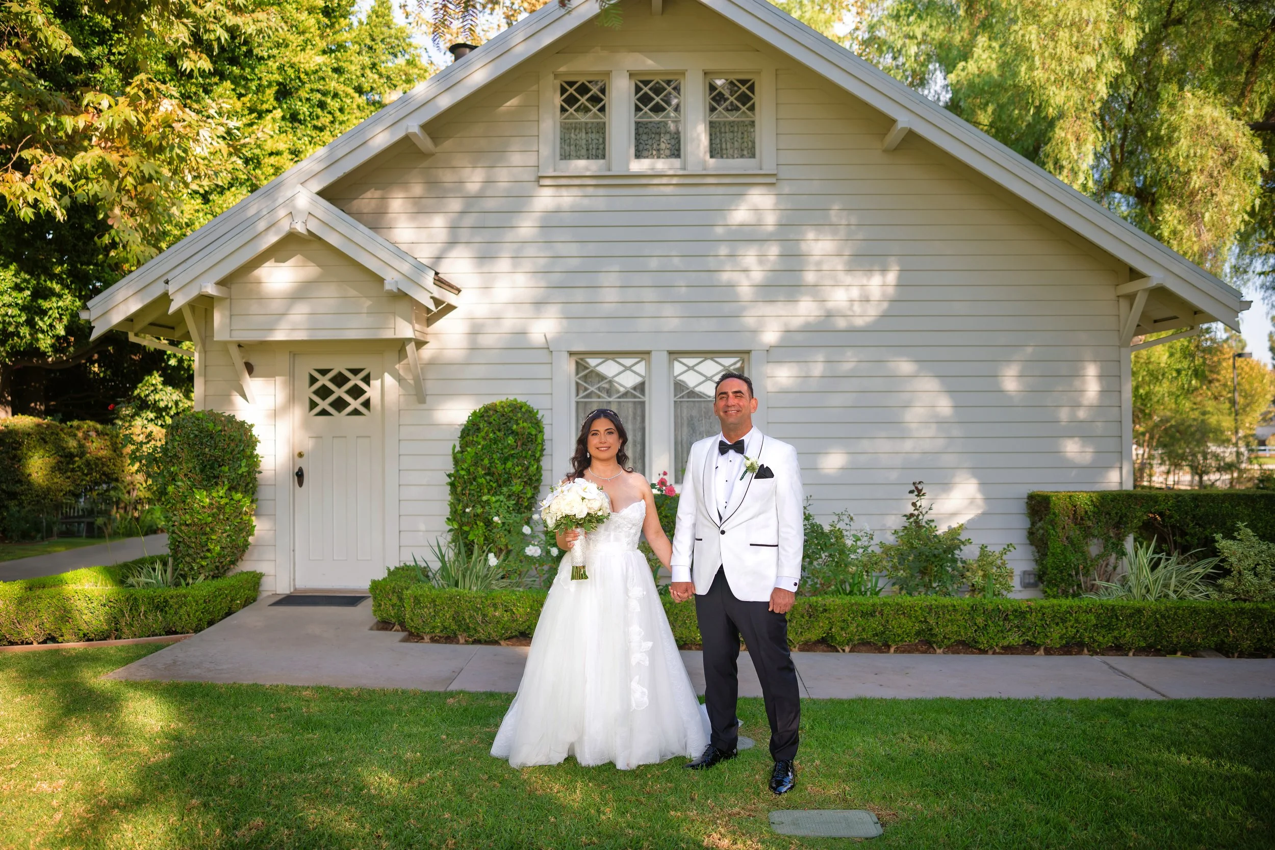 A bride and groom holding hands and smiling in front of a white house with greenery, dressed in wedding attire, with the bride holding a bouquet of white flowers.