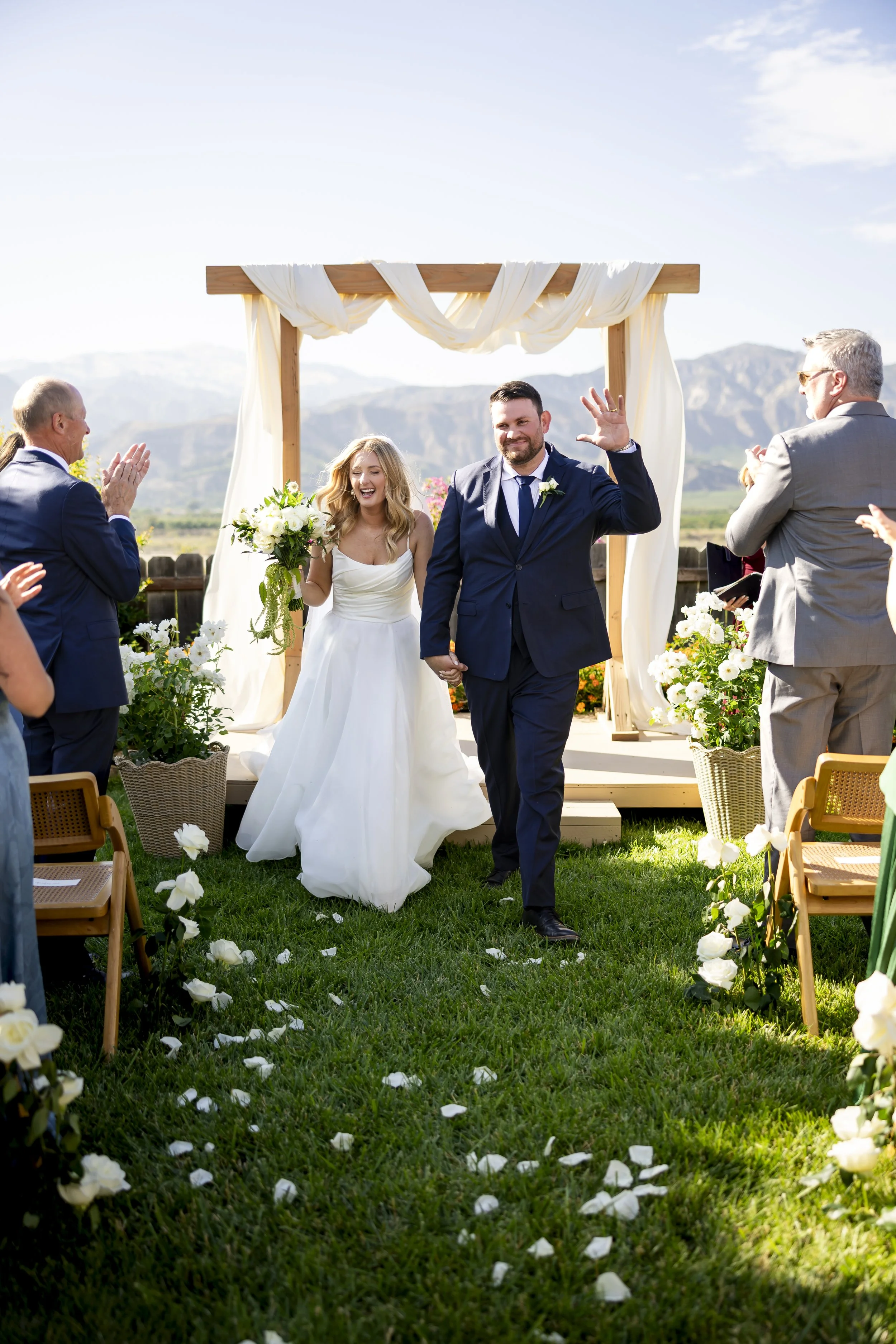 A newlywed couple walking hand-in-hand down an outdoor aisle, smiling and waving at their guests during a wedding ceremony against a scenic mountain backdrop, with flower petals on the grass.