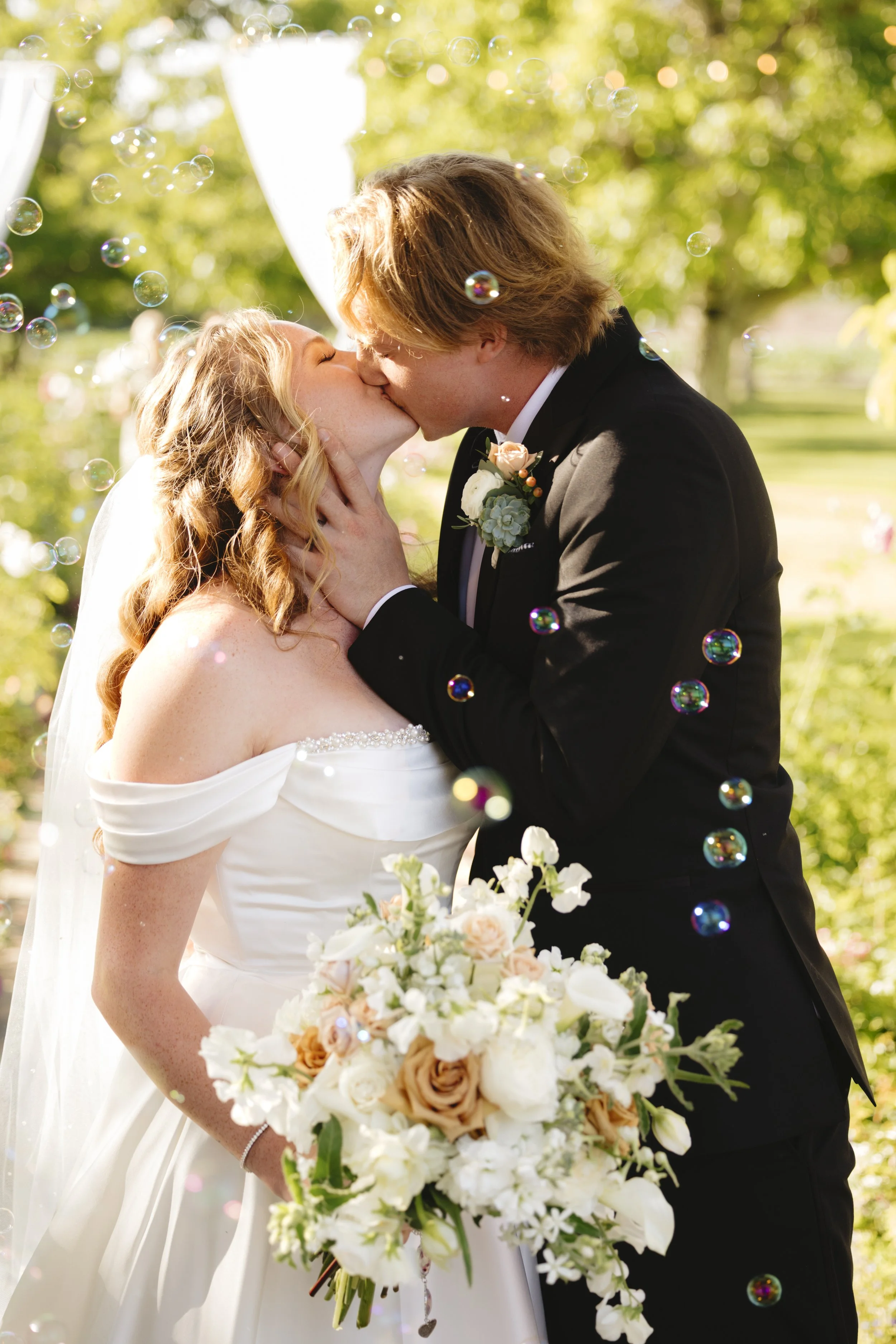 A bride and groom kiss outdoors during their wedding, with bubbles floating around them and a lush green background.