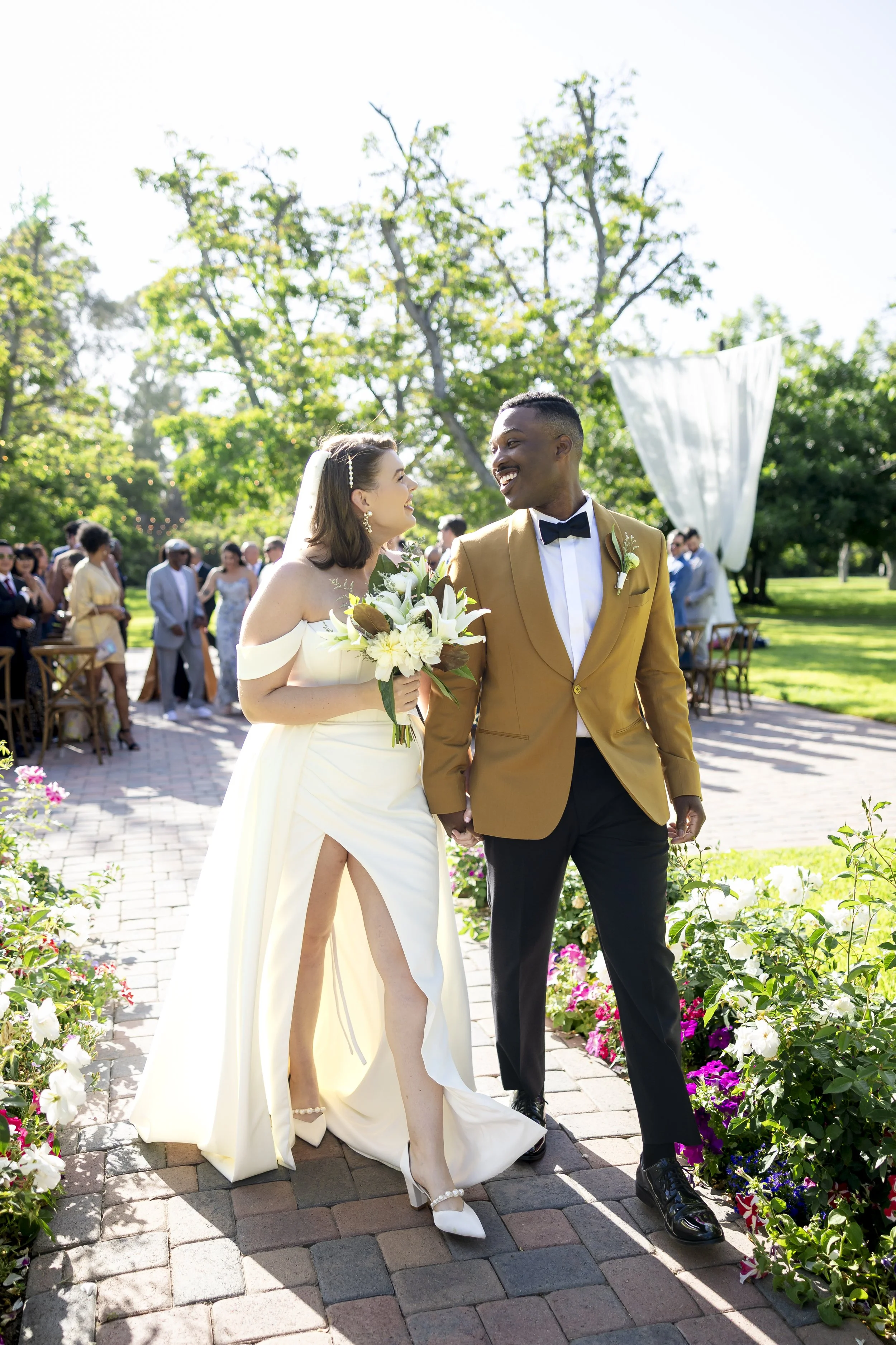 Bride and groom holding hands and smiling during their outdoor wedding ceremony, with guests in the background and decorated greenery.