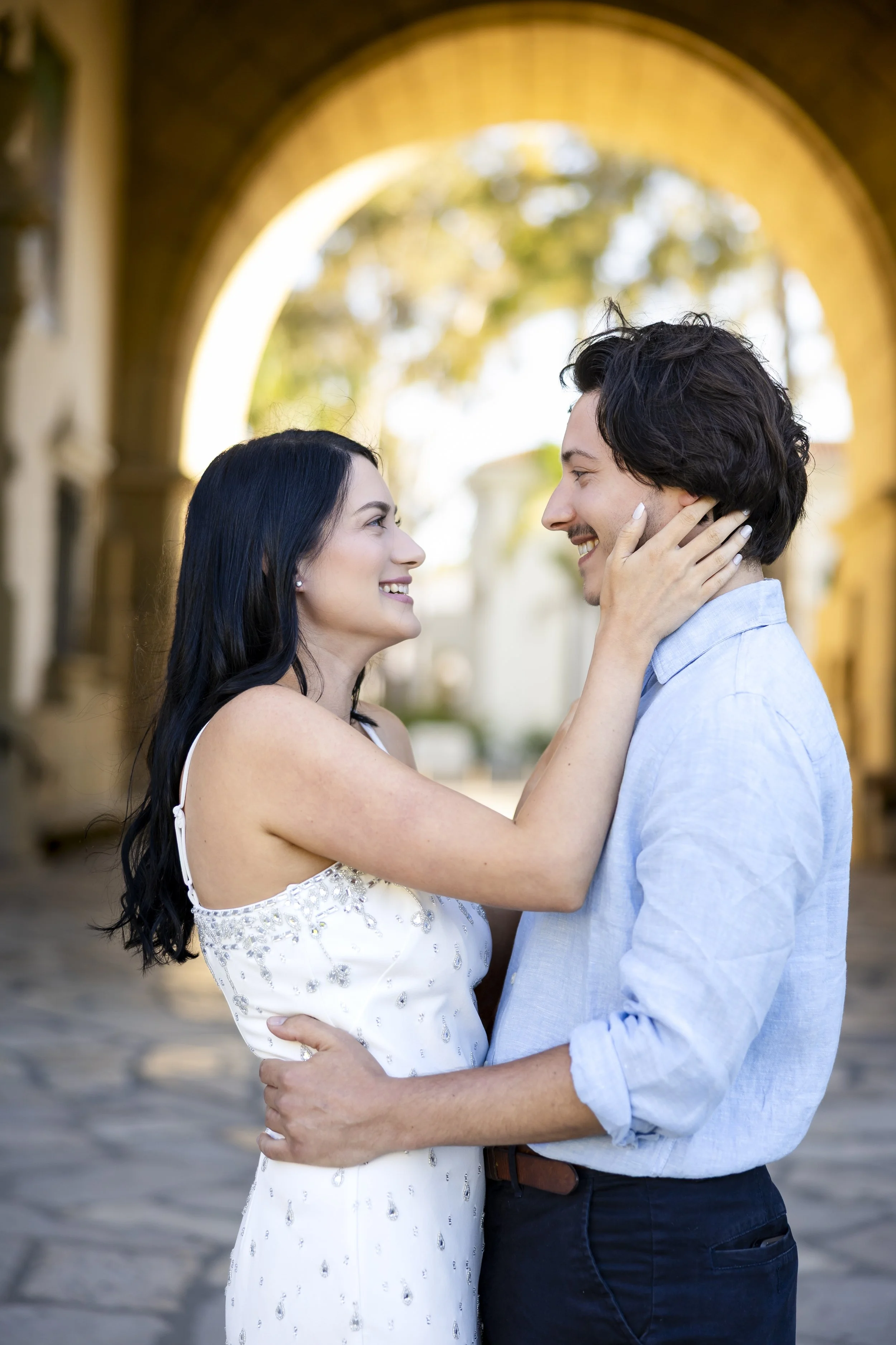 A happy couple in an embrace, smiling at each other, standing under an arched stone structure outdoors.
