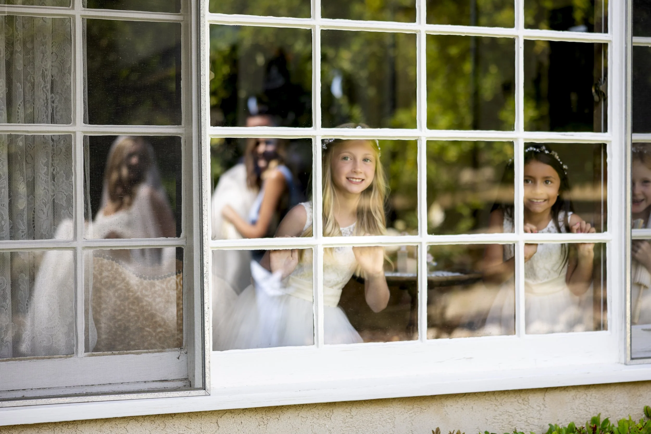 Children looking out of a window with a grid pattern, smiling and dressed in white dresses, outdoors with green trees in the background.