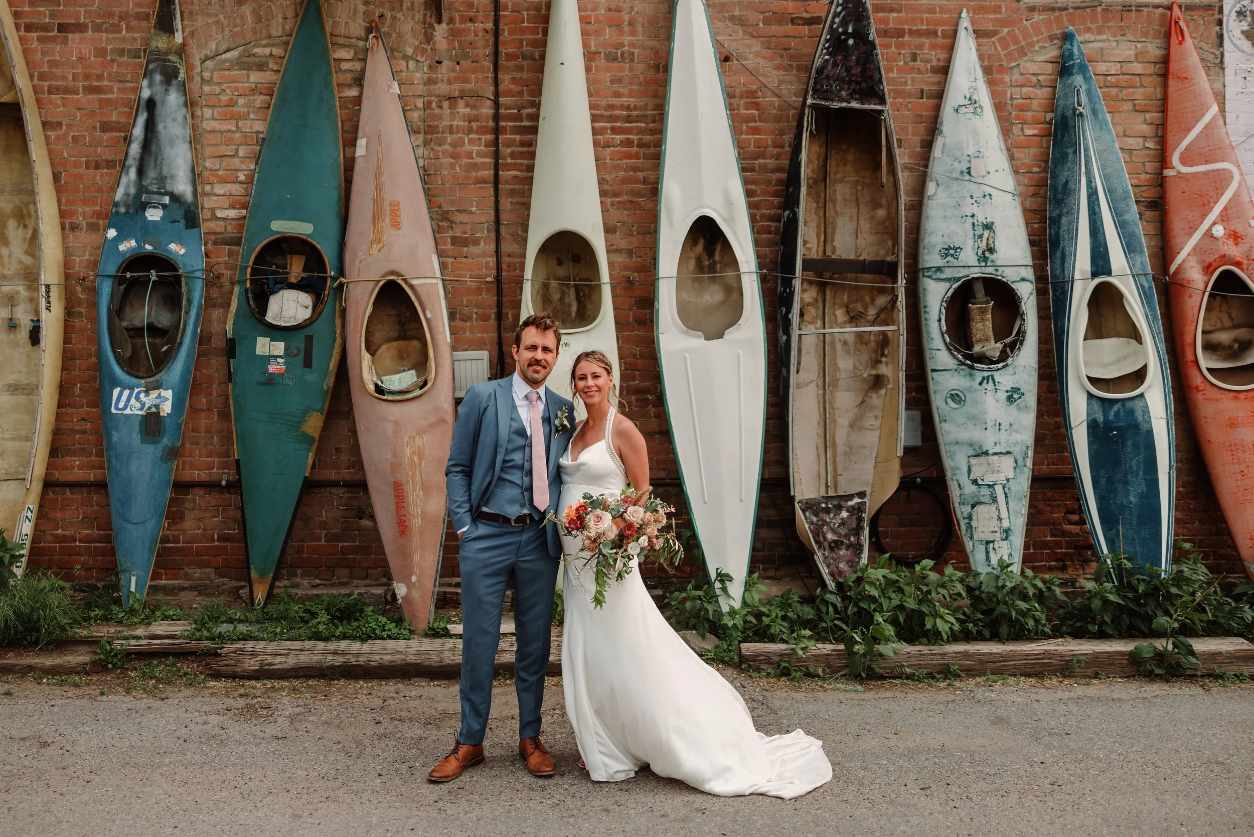 A newlywed couple standing in front of a brick wall decorated with old, colorful boats. The bride is holding a bouquet and the groom is wearing a light blue suit.