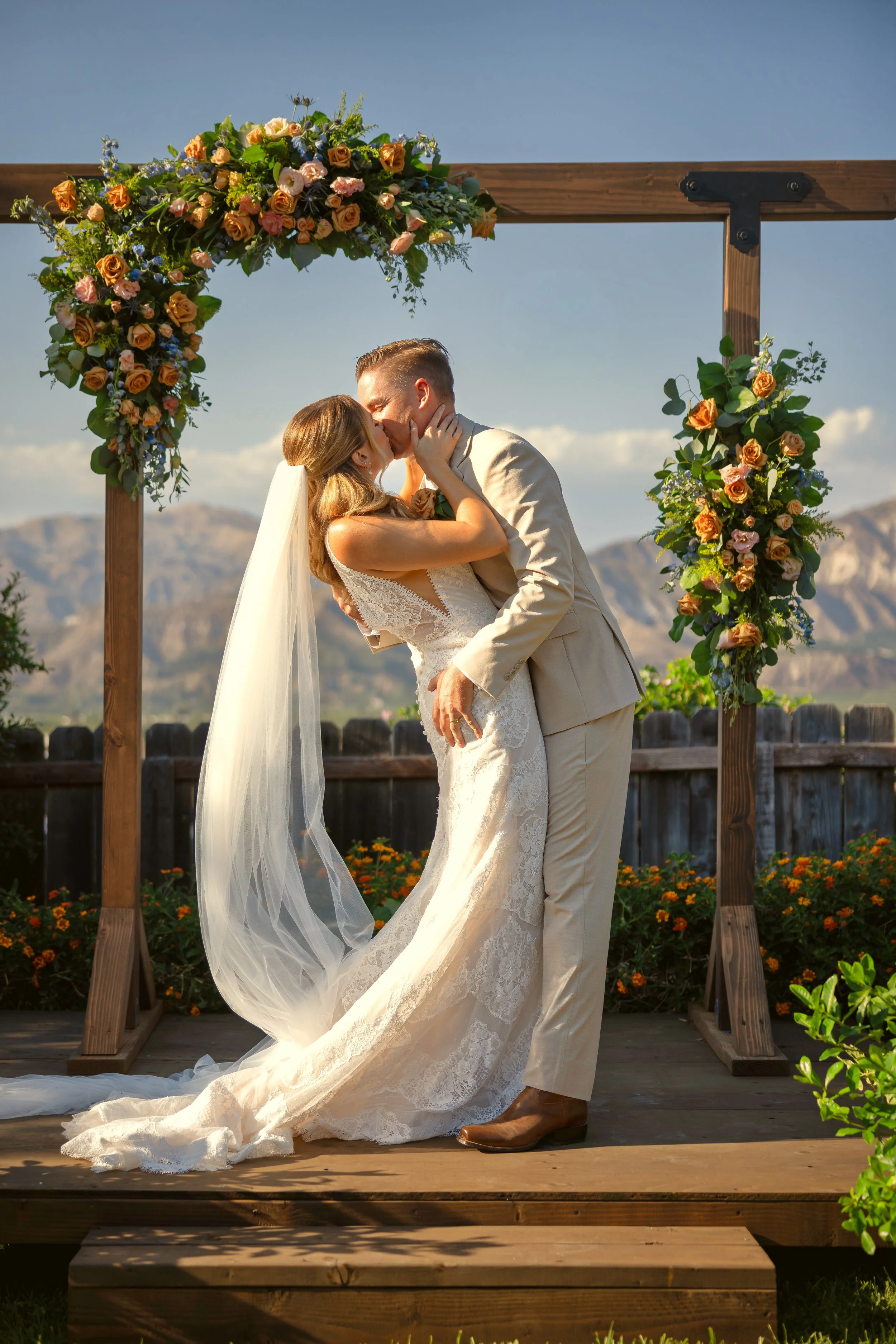 A bride and groom share a kiss at their outdoor wedding ceremony under a floral arch with mountains in the background.