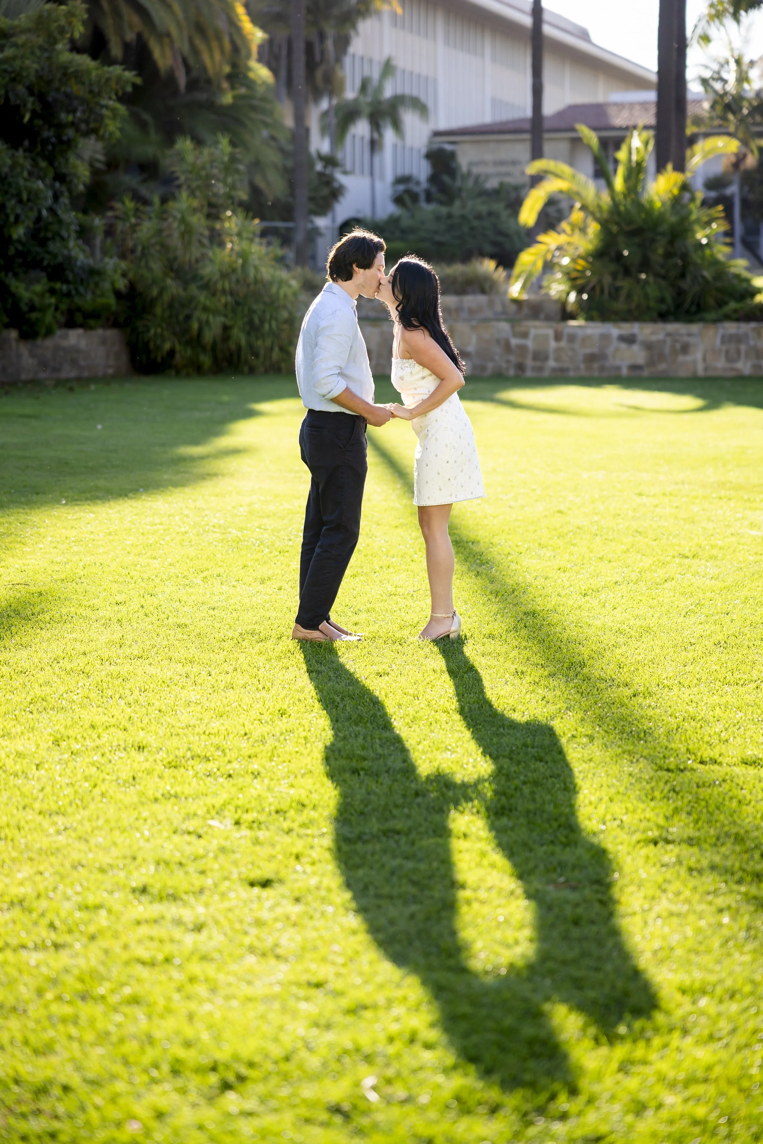 A couple holding hands and kissing on a sunny, grassy lawn.