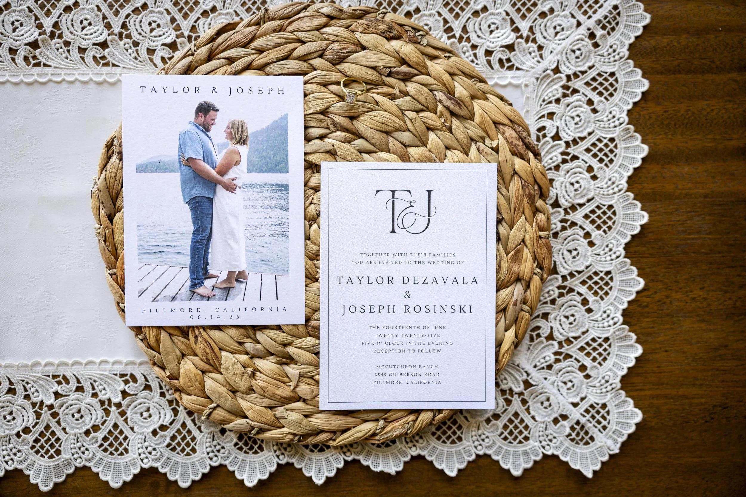 Wedding invitation and photo card on a woven placemat, with lace doily underneath, on a wooden surface. The invitation has initials 'T & J', names 'Taylor Dezavala & Joseph Rosinski', and details of the wedding at McCutcheon Ranch in California.