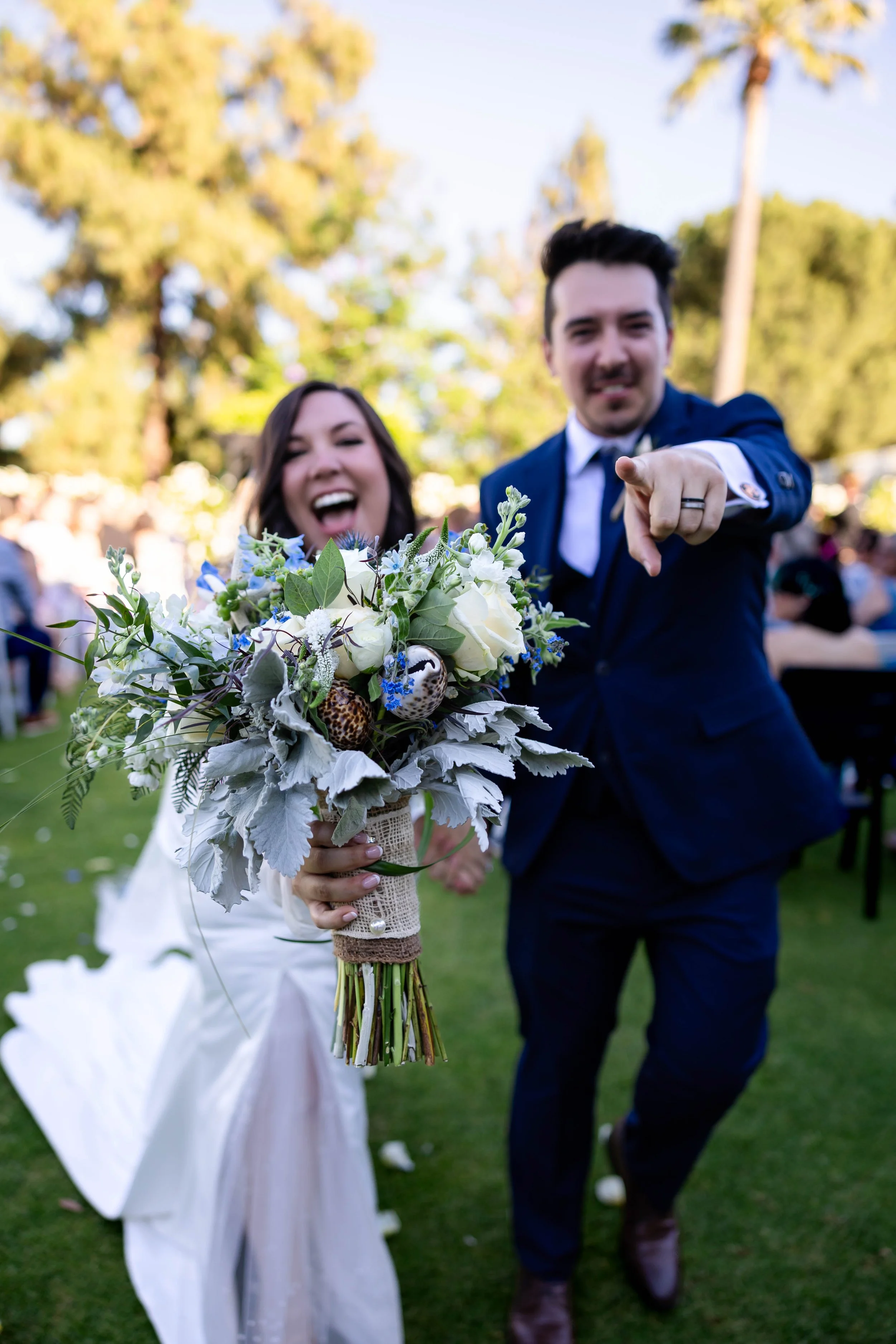 Happy bride and groom at an outdoor wedding, with the bride holding a large bouquet of white and purple flowers, and the groom pointing at the camera.