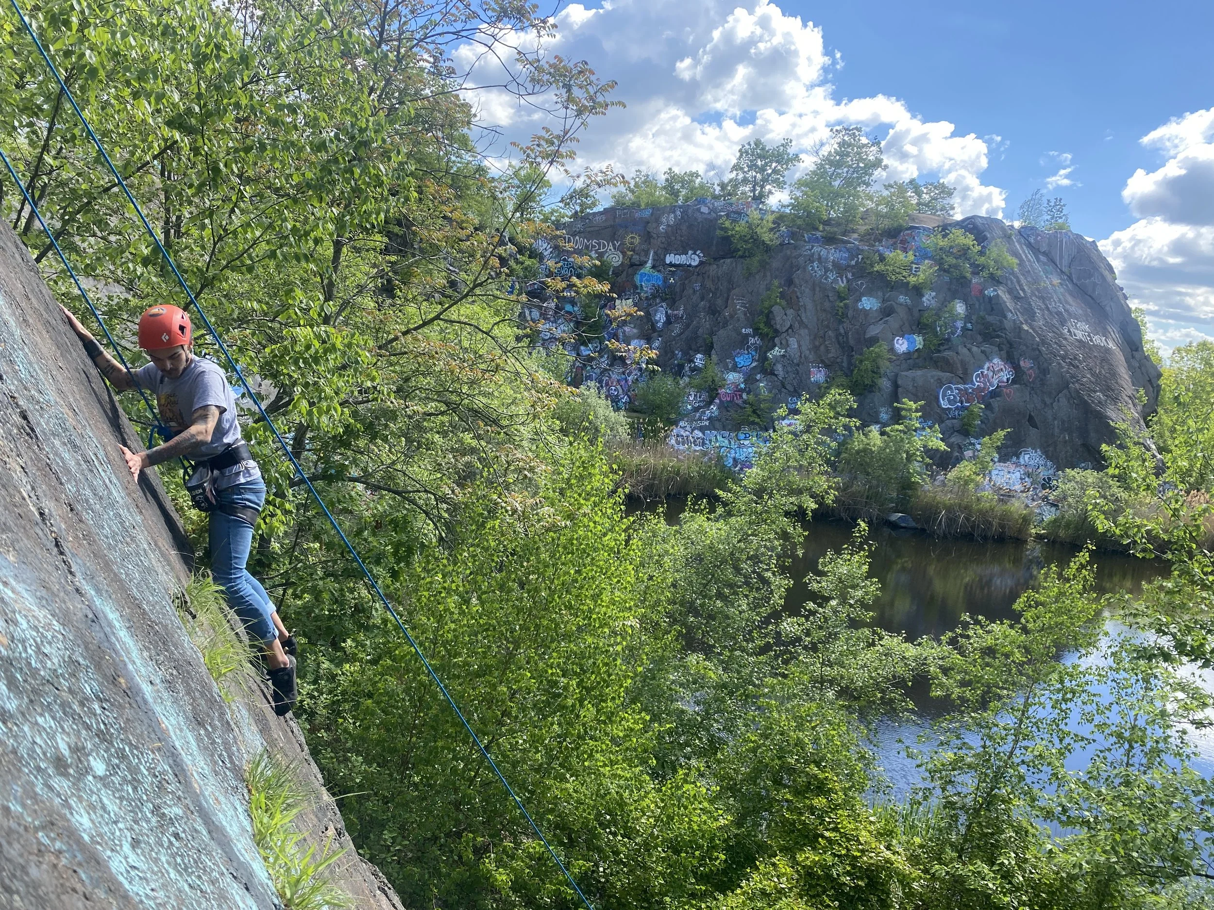 Climber scales a wall at Quincy Quarry with a pond and grafitti covered cliffs in the background