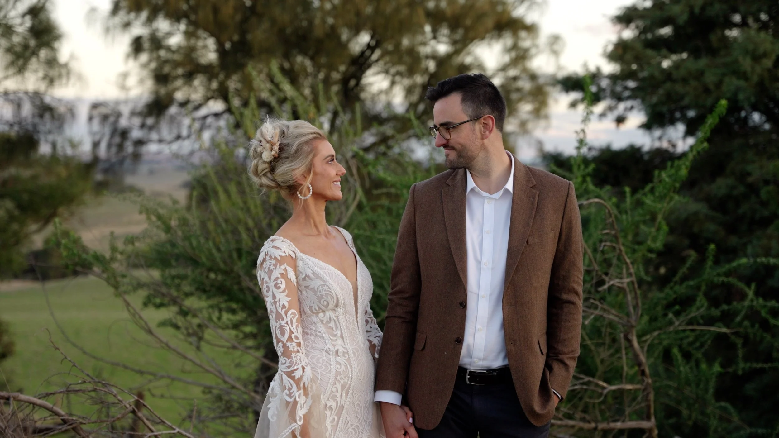 A bride and groom are standing outdoors, holding hands and looking at each other with smiles. The bride is wearing a white lace wedding dress and the groom is wearing a brown jacket with a white shirt. There are trees and bushes in the background.