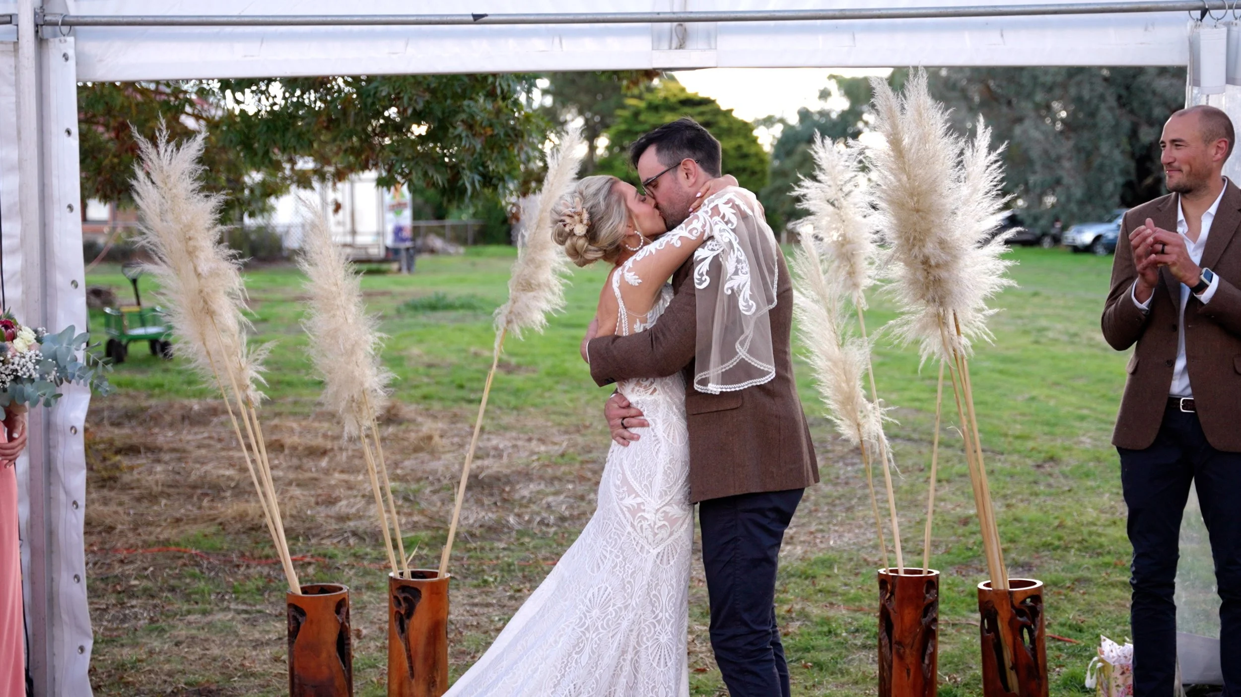 Bride and groom hugging during their outdoor wedding ceremony with decorative pampas grass in wooden vases.