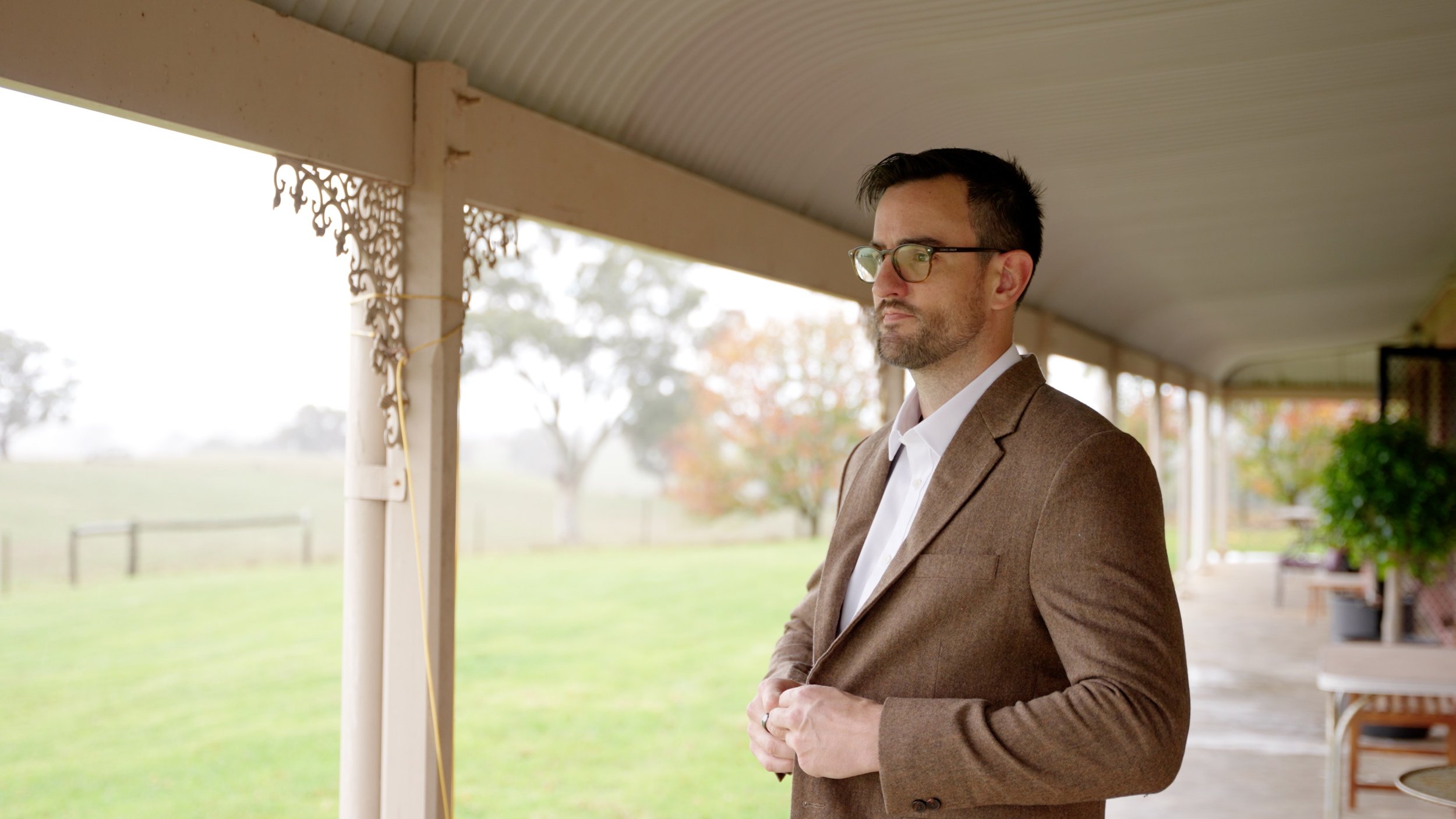 A man with dark hair, glasses, wearing a brown blazer and white shirt, standing on a porch with a grassy field and trees in the background.