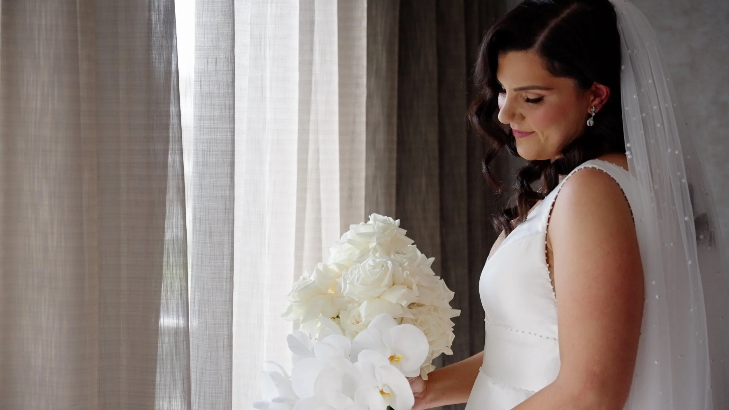 Bride in a white wedding dress holding a bouquet of white flowers, standing near a window with sheer curtains, smiling softly.